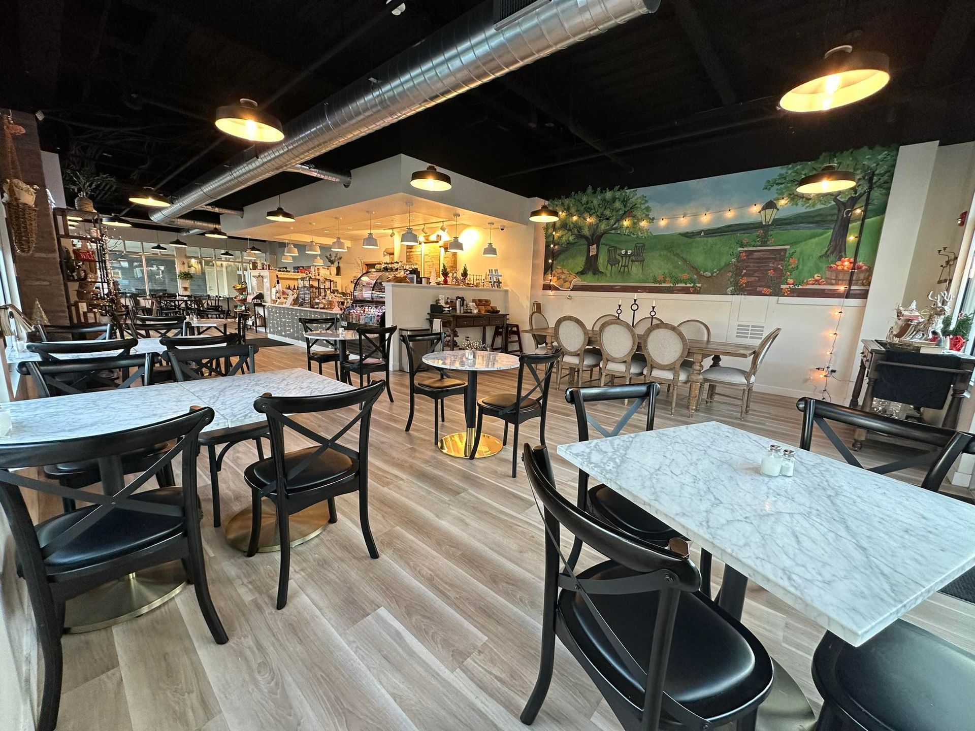 Interior view of a cozy cafe with light wood floors, marble-topped tables, black cross-back chairs, and a painted mural.