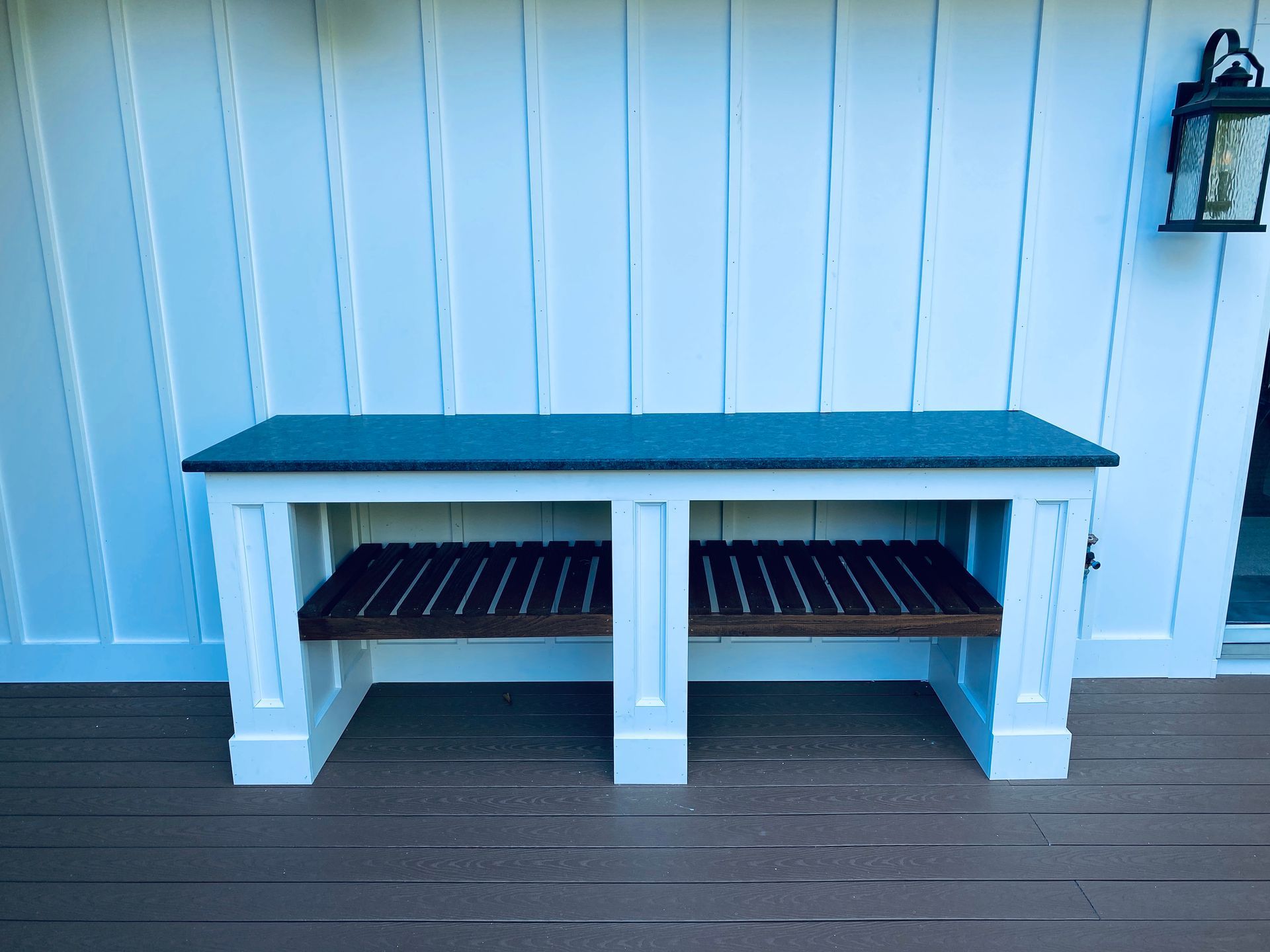 A wooden bench is sitting on a wooden deck in front of a white wall.