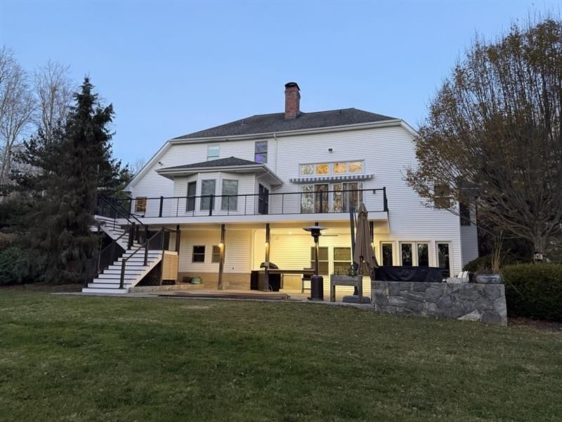 A white, two-story house with a large upper deck and a ground-level patio illuminated by warm lights at twilight.