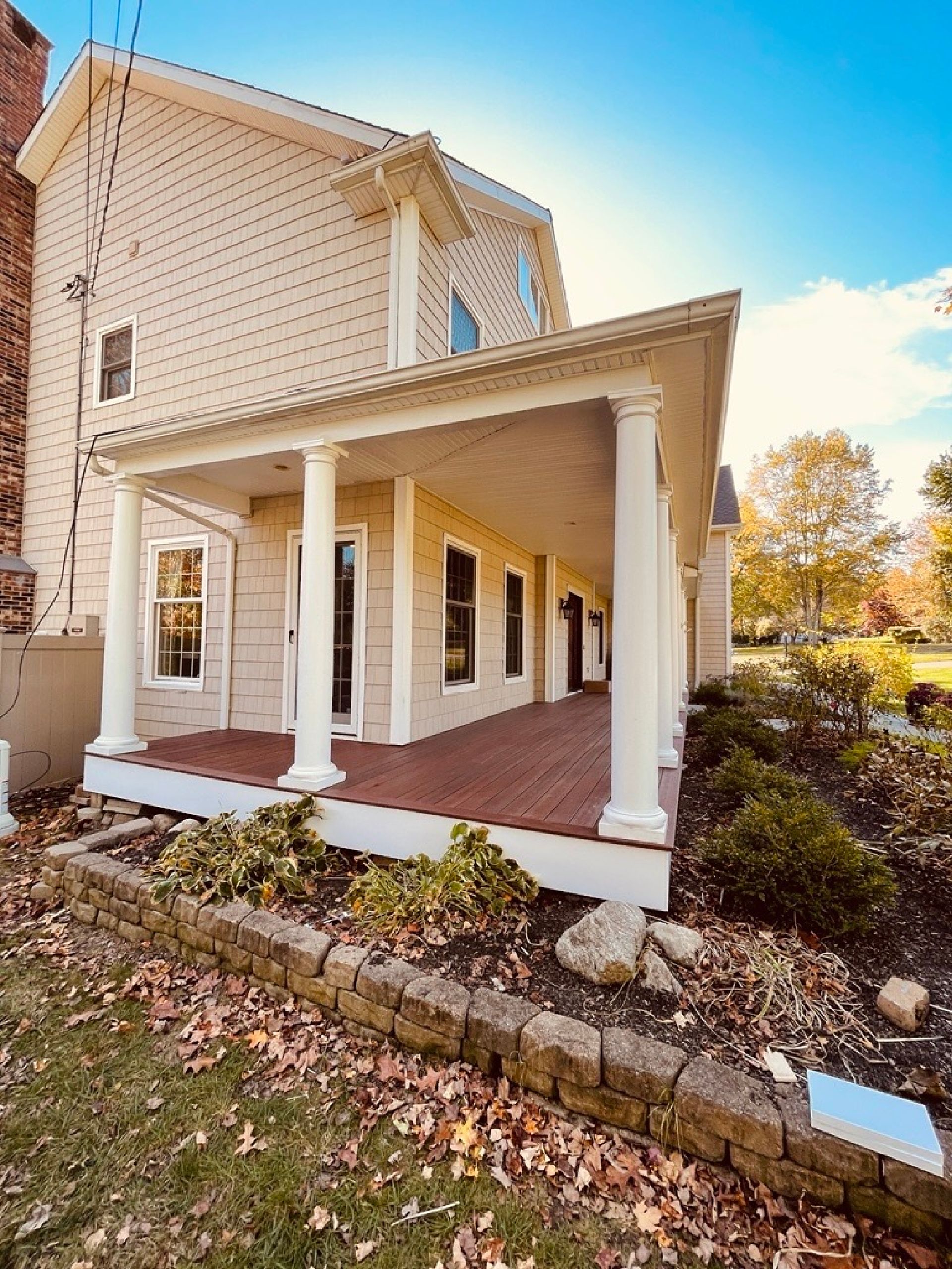 Tan-sided house with a large wrap-around porch featuring white columns and a reddish-brown deck, set in a yard with trees.