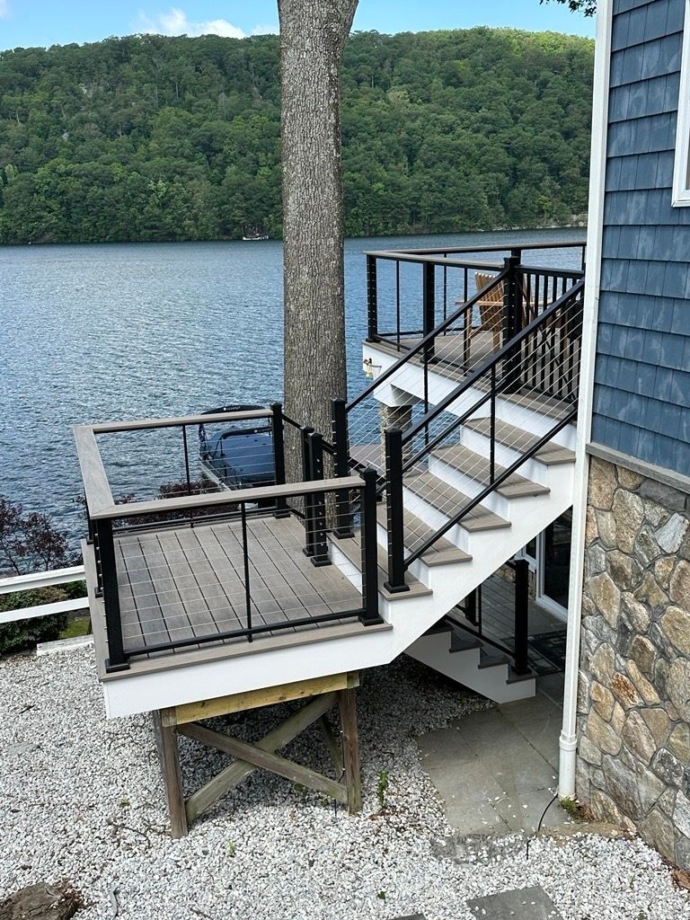 A tiered wooden deck with black railings leads down to a pebble-covered lakeside yard beside a shingled house.