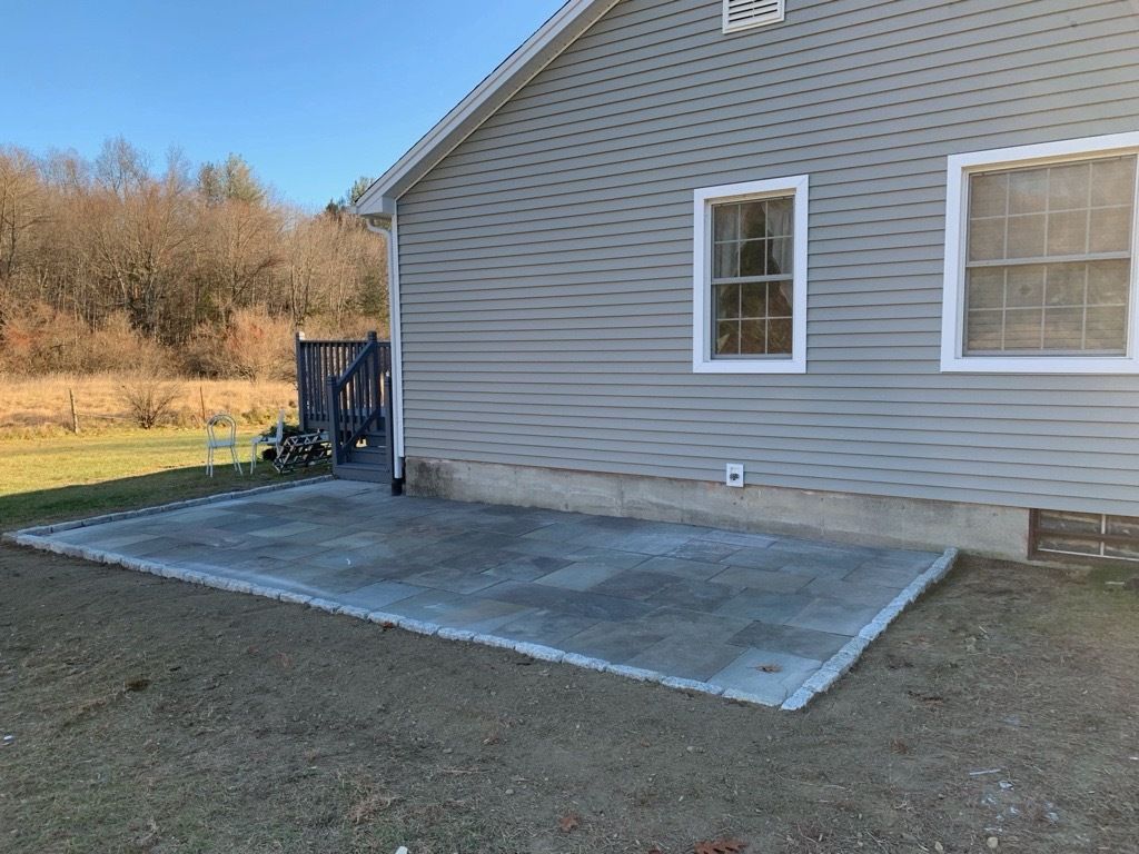 A rectangular concrete patio borders a gray-sided house with two windows and a concrete foundation on a sunny day.