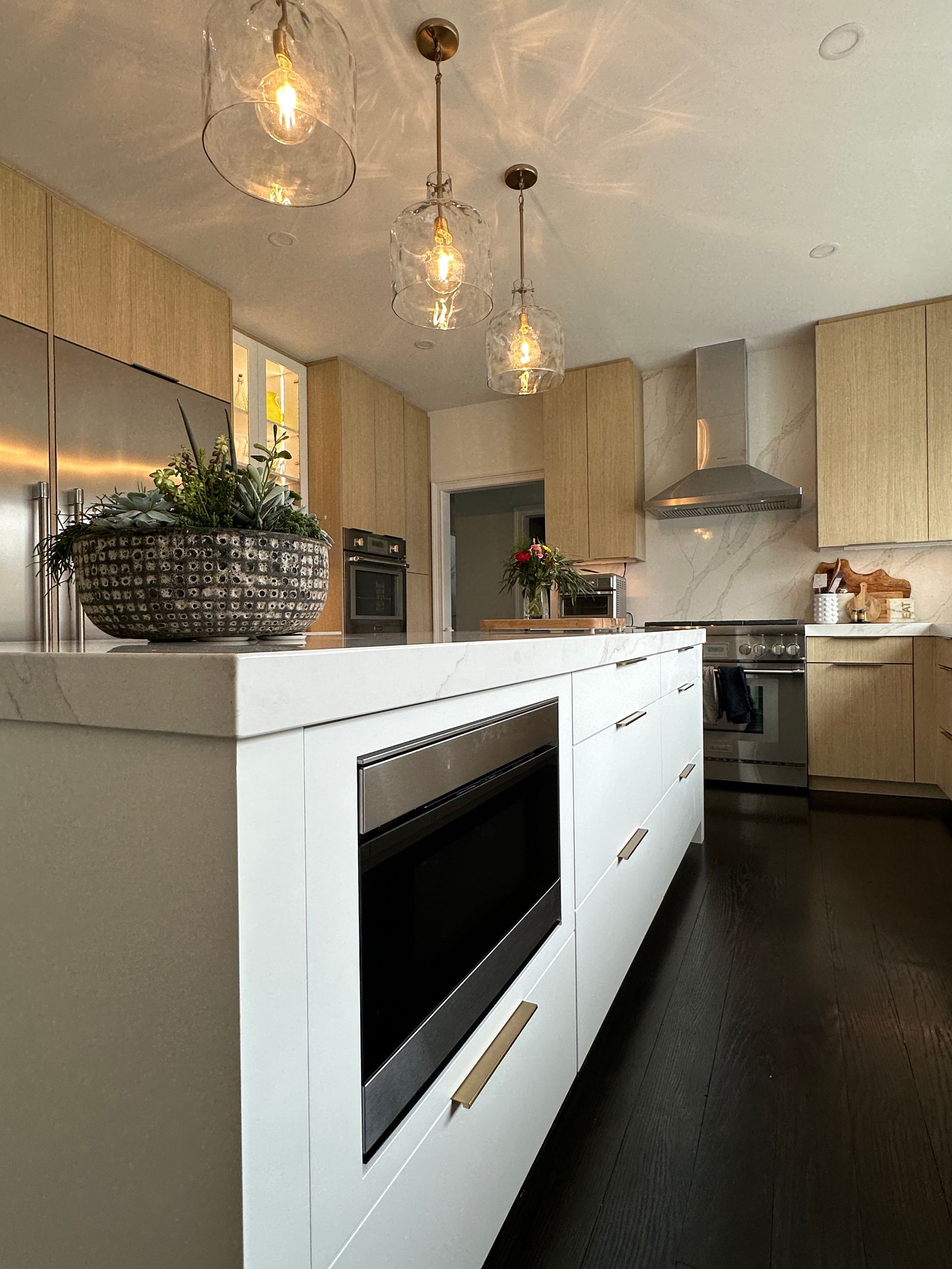 Modern kitchen with a white island, light wood cabinets, three pendant lights, stainless steel appliances, and dark floors.