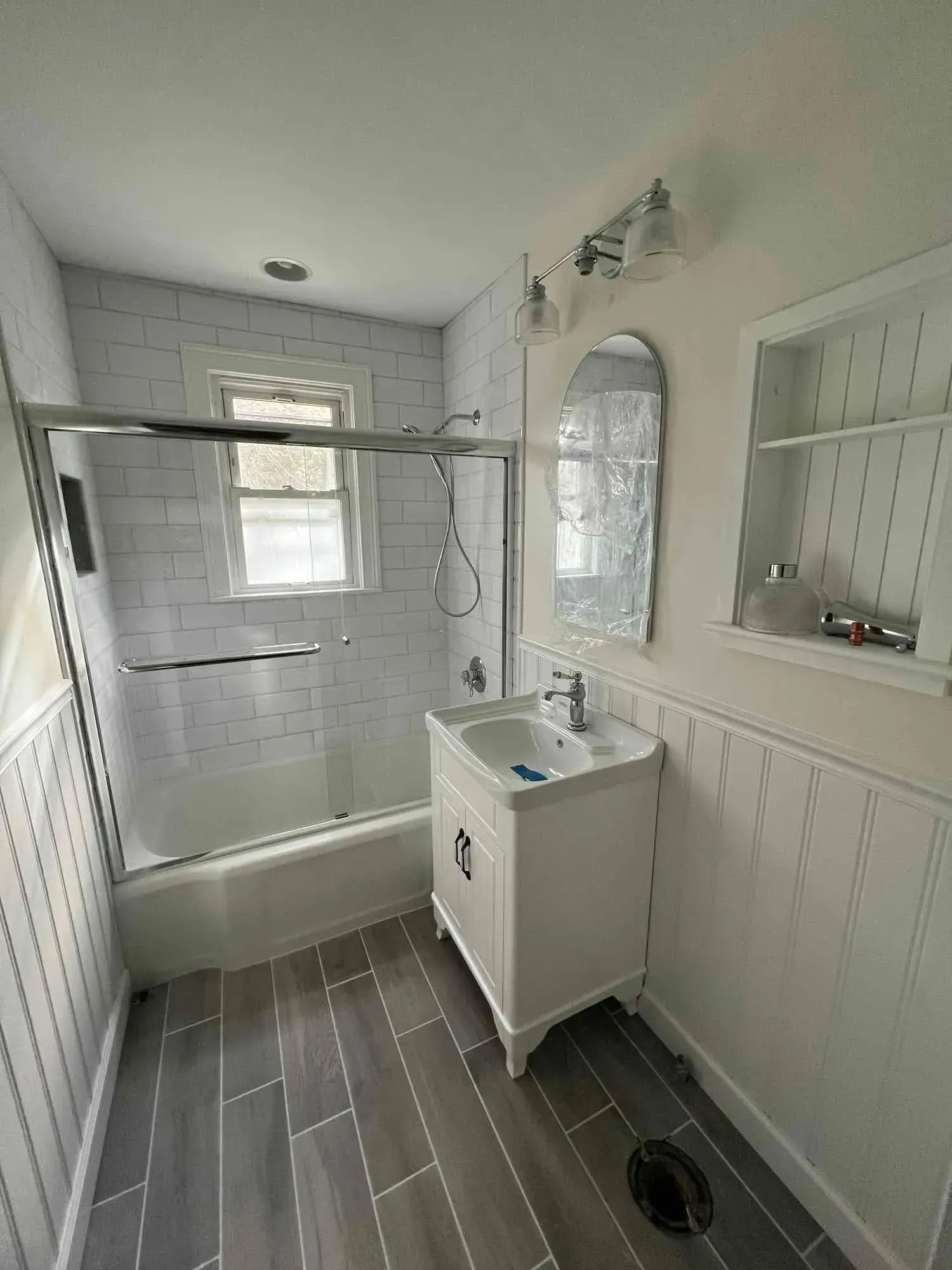 A renovated bathroom with a white vanity, a tiled bathtub-shower combination, gray wood-look floors, and built-in shelving.