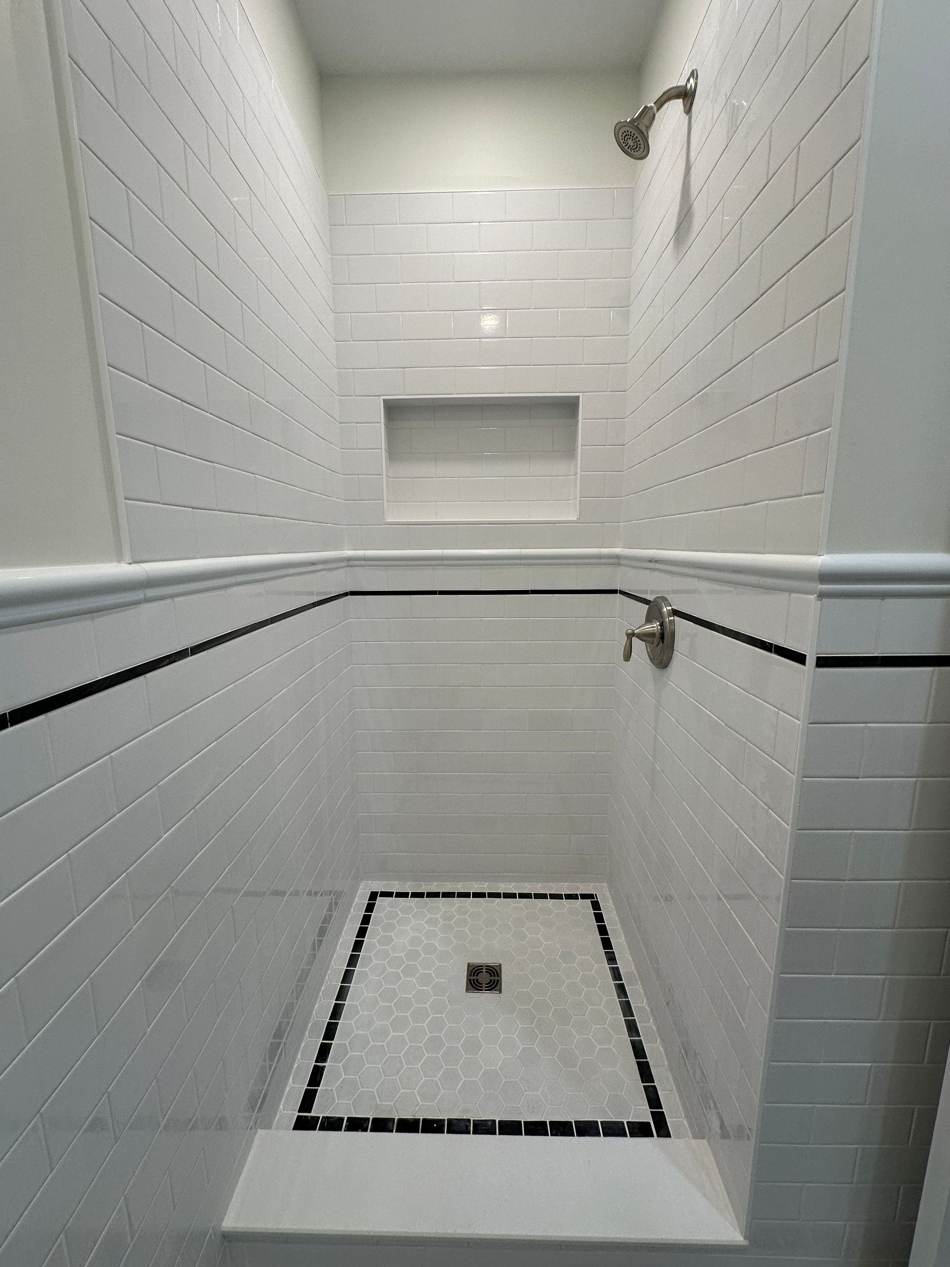A white-tiled shower with black accent trim, a built-in recessed shelf, and a square central drain.
