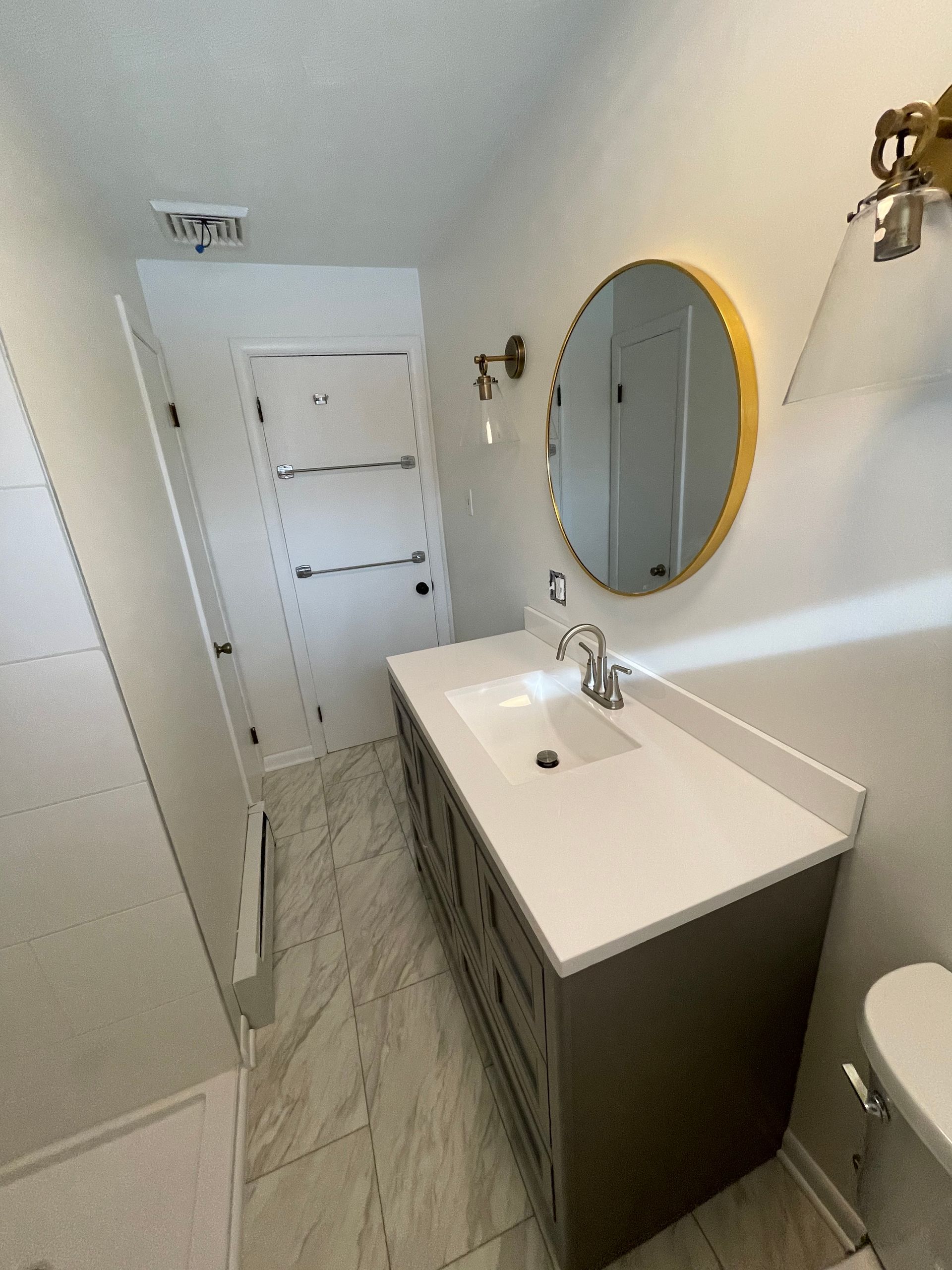 A modern bathroom with a grey vanity, white quartz countertop, round mirror, and marble-style tile floors.