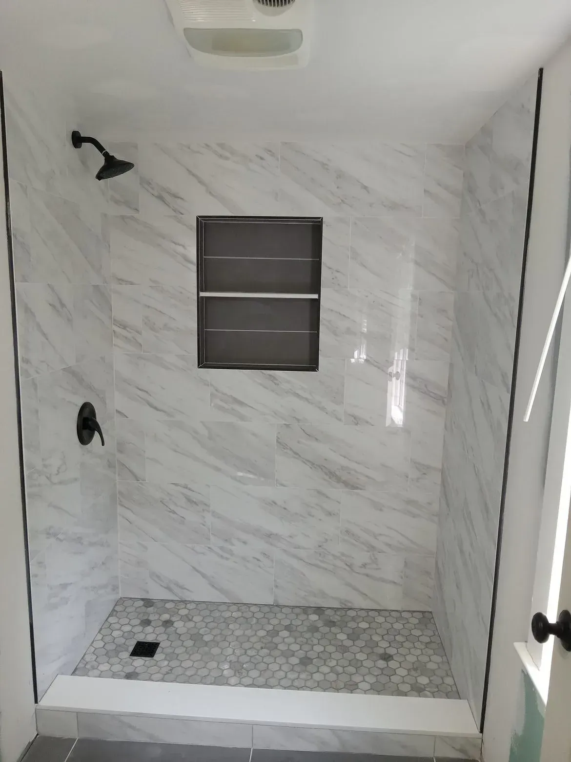 A walk-in shower with white marble-patterned tile walls, a gray mosaic tile floor, and black matte fixtures.