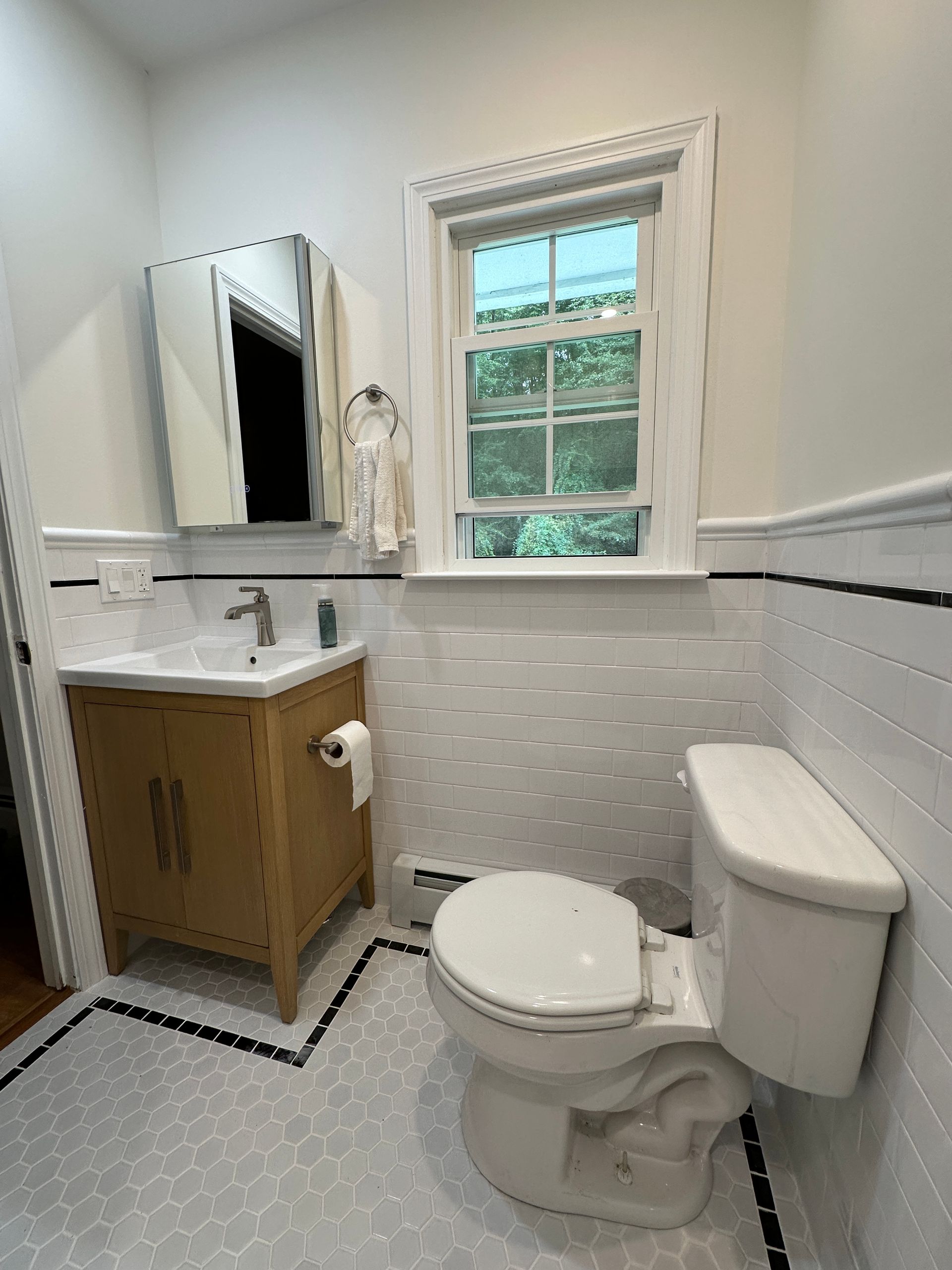 A small bathroom with a wooden vanity, a white toilet, subway tile walls, and a patterned tile floor beneath a window.