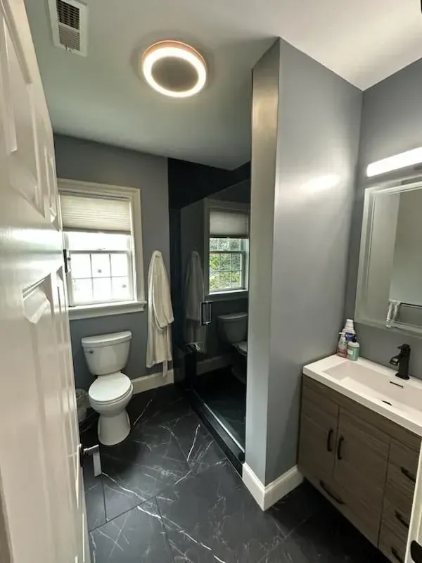 A modern bathroom with grey walls, dark marble-look tile floors, a white toilet, a walk-in shower, and a wood vanity.