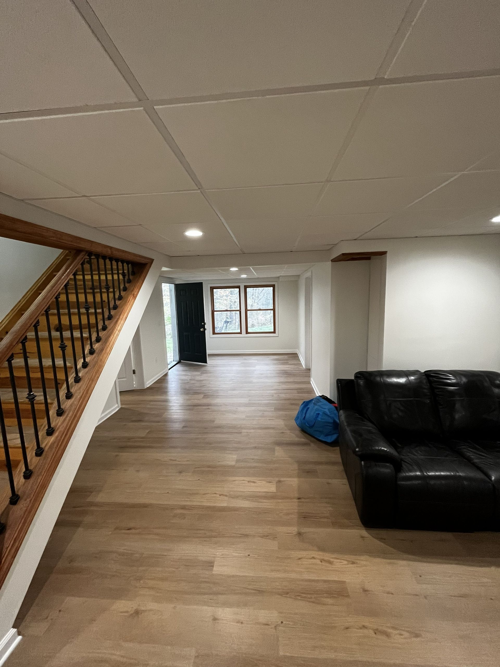 A finished basement with light wood flooring, a staircase with wooden railings, and a black leather sofa on the right.