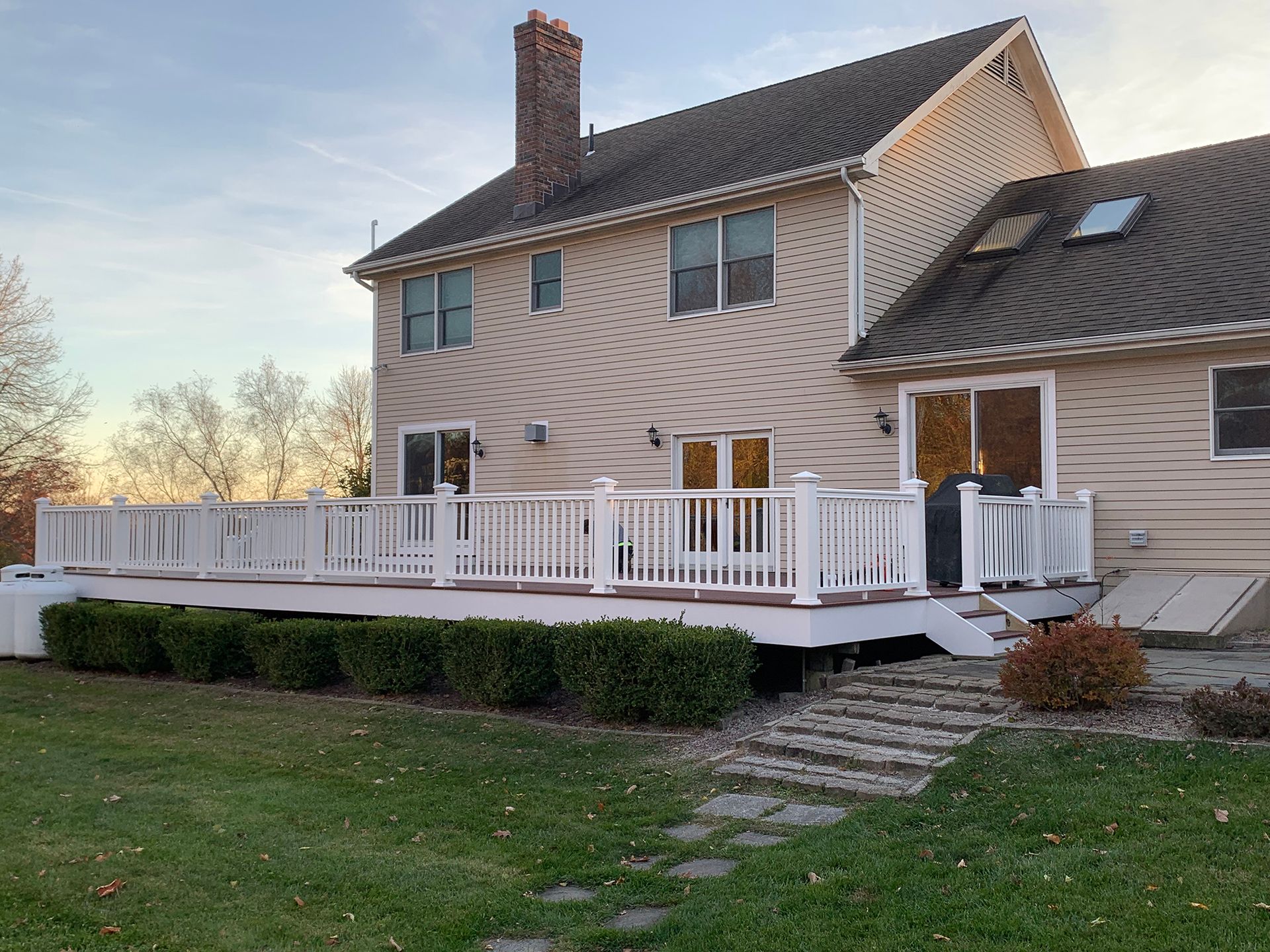 A two-story tan house with a large white deck, brick chimney, and a stone path leading to a grassy lawn at dusk.