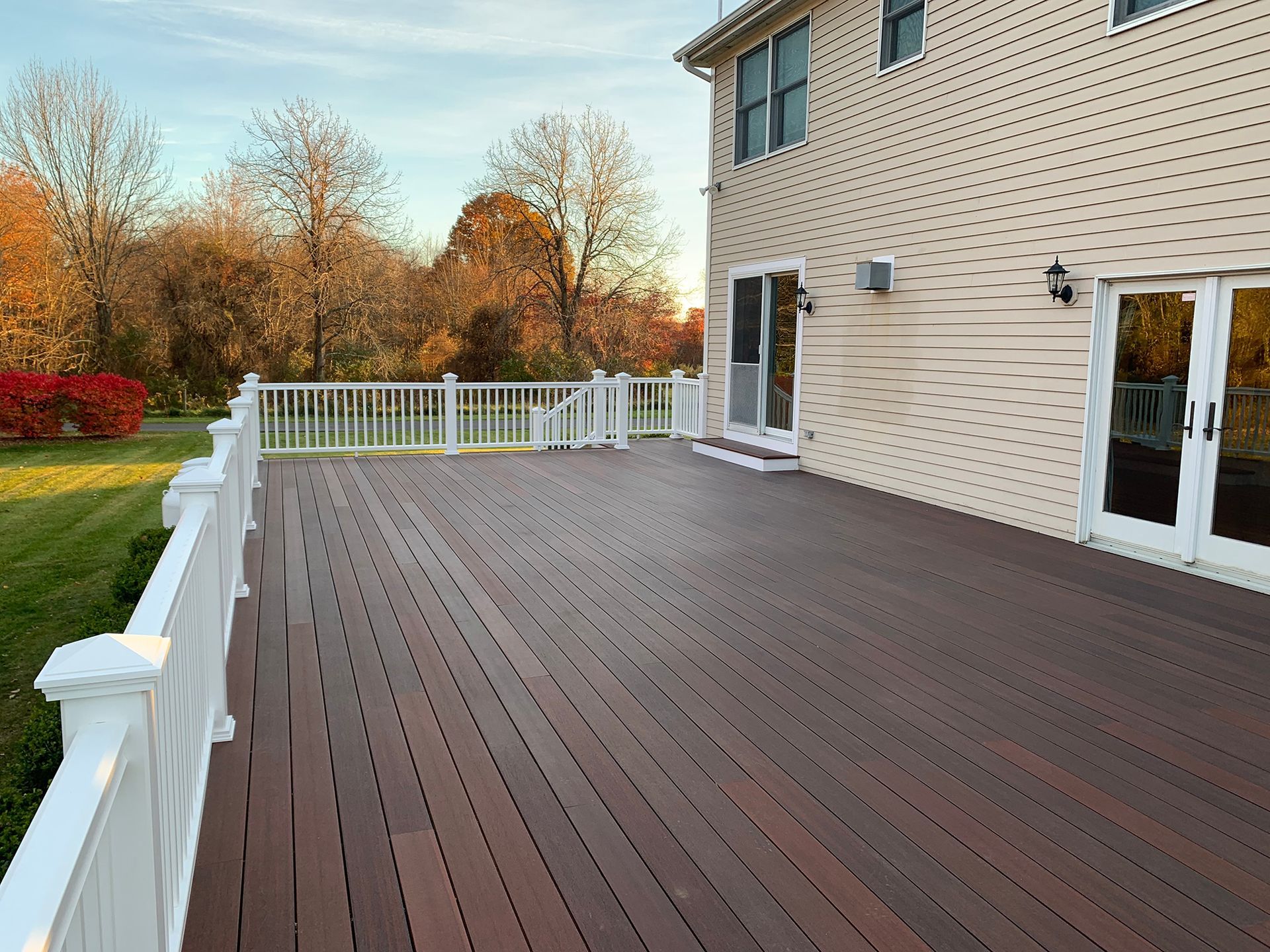 A wide, dark brown wooden deck with white railings attached to the side of a beige two-story house near a lawn.