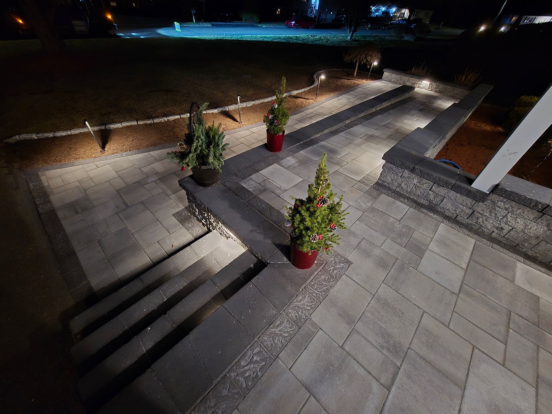 Night view of a stone patio with steps, illuminated by small path lights and featuring two small potted evergreen trees.