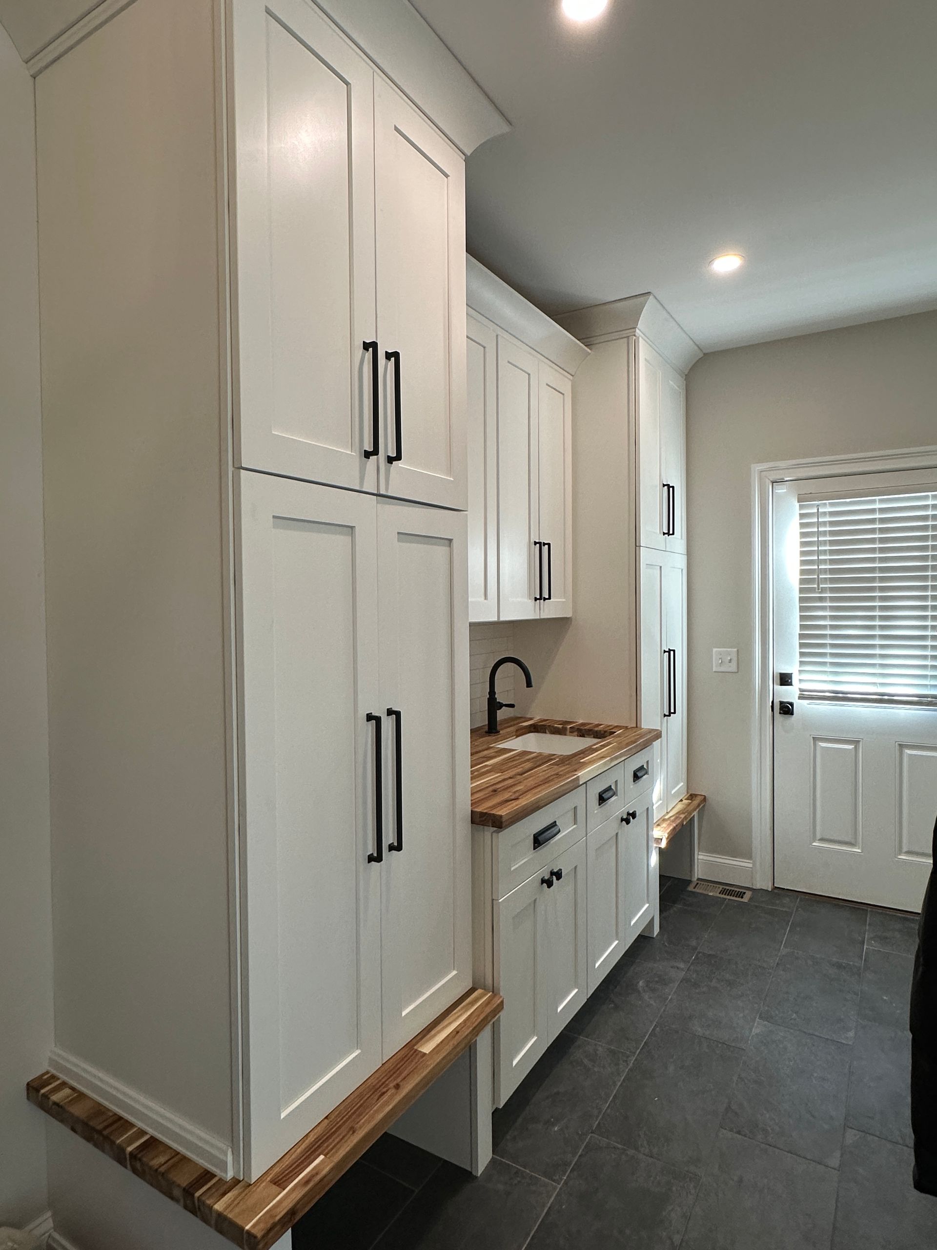 White cabinets with black hardware and wood countertops in a kitchen or mudroom area with a door in the background.