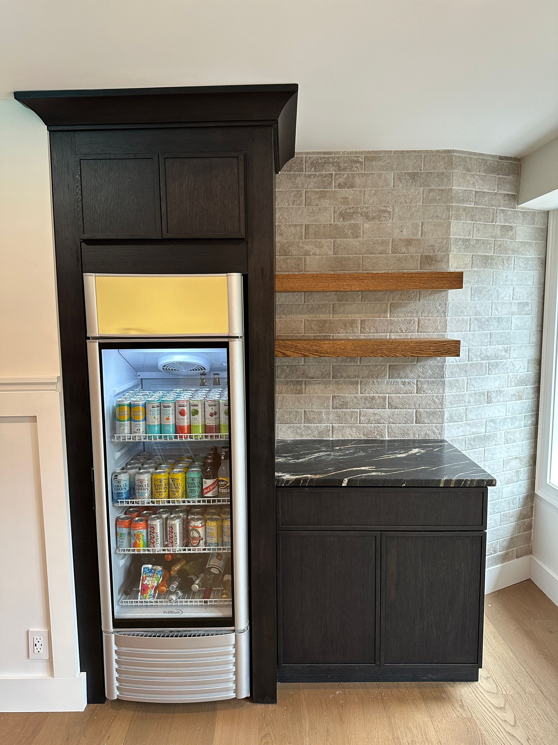 A black wood wet bar with a glass-front beverage fridge, stone tile backsplash, floating wooden shelves, and dark counter.