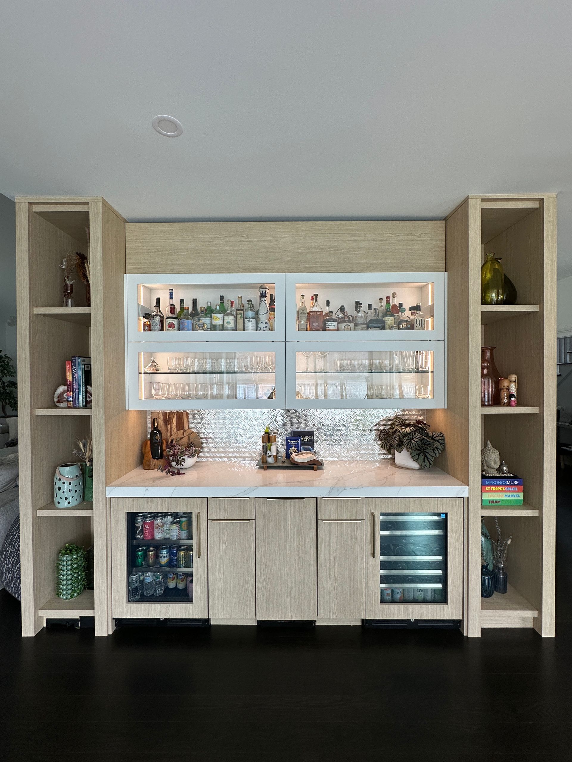 A home bar built into a wall, featuring light wood cabinetry, white countertops, glass shelves, and a beverage fridge.