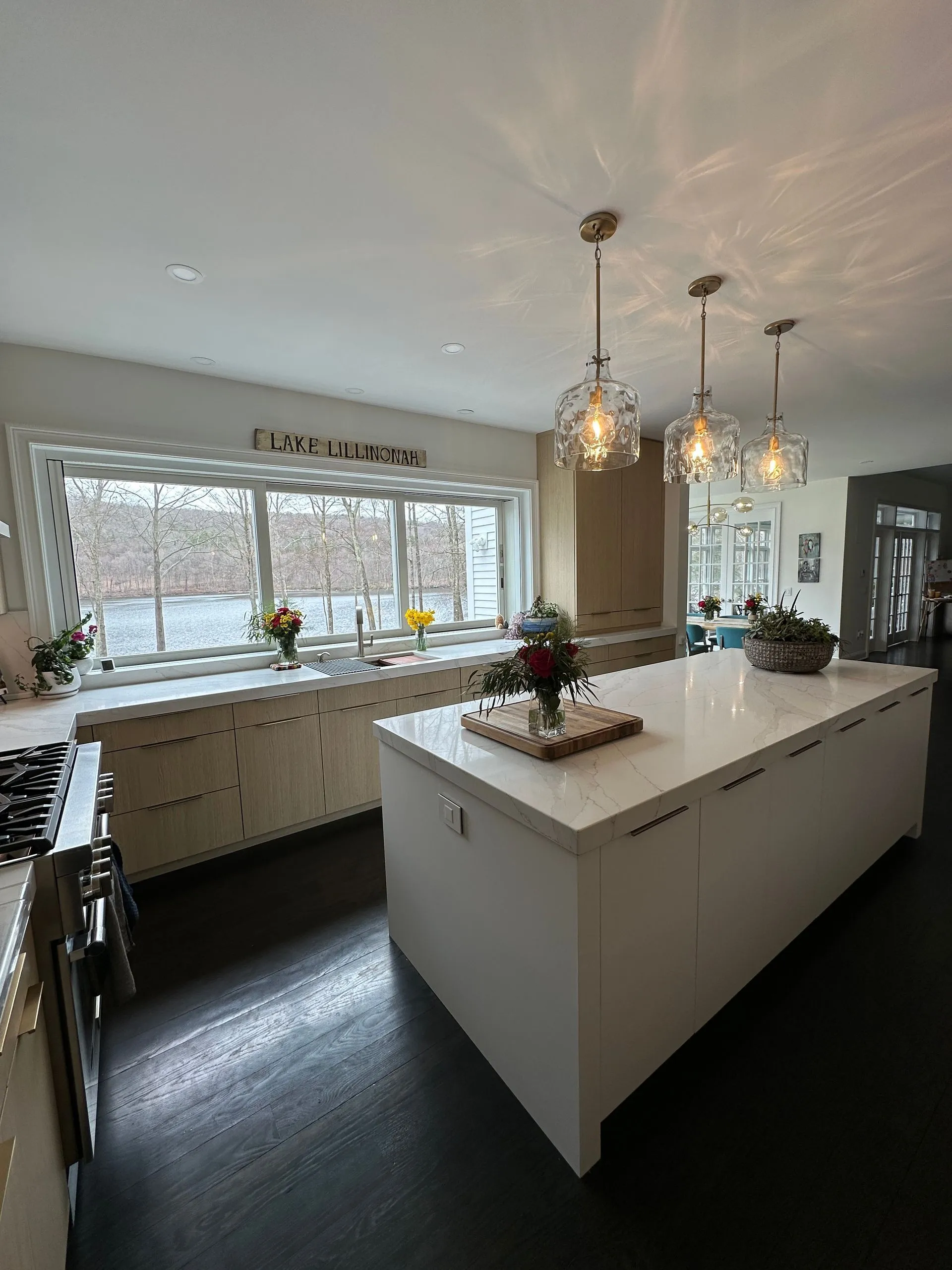 A modern kitchen featuring a large white island, wooden cabinets, and three glass pendant lights under a windowed view.