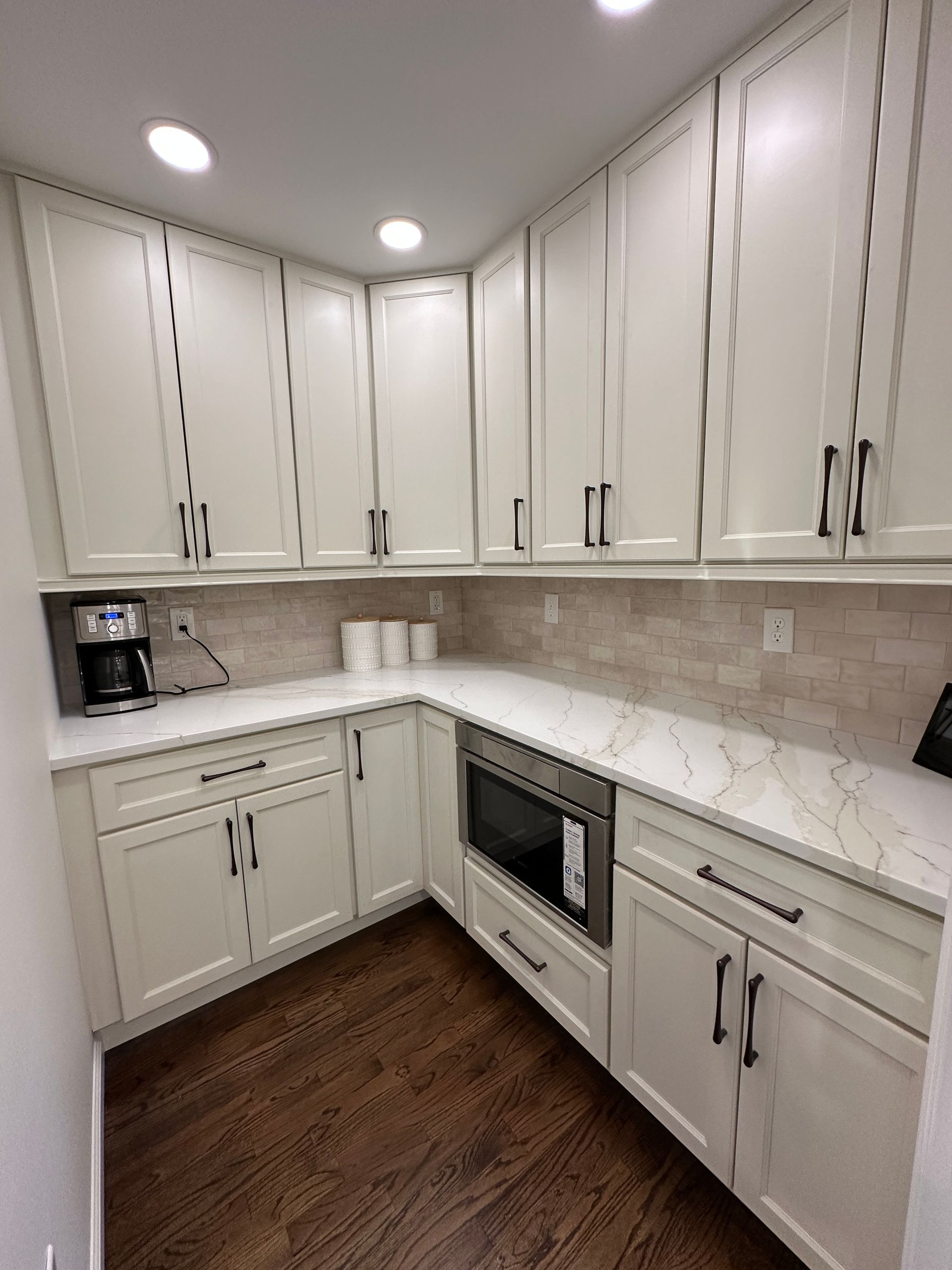 A kitchen corner featuring white shaker cabinets, white marble countertops, a subway tile backsplash, and dark wood floors.