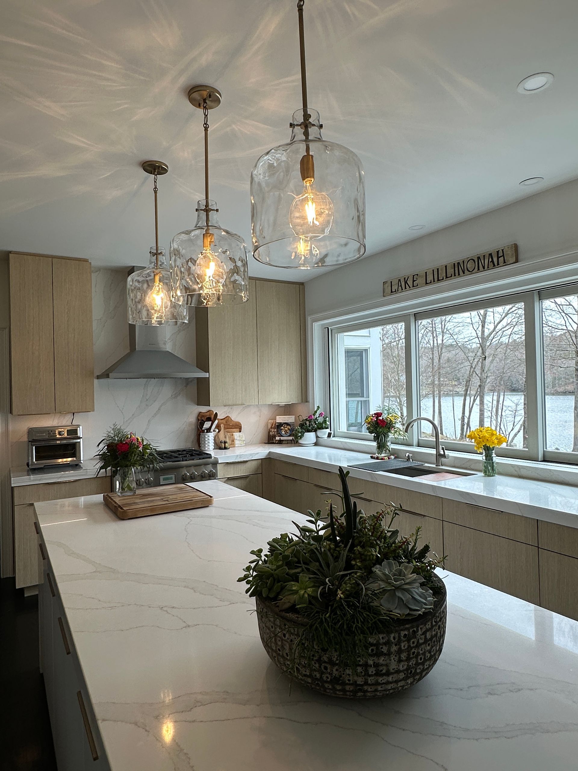 A modern, bright kitchen with light wood cabinets, white countertops, an island centerpiece, and three glass pendant lights.