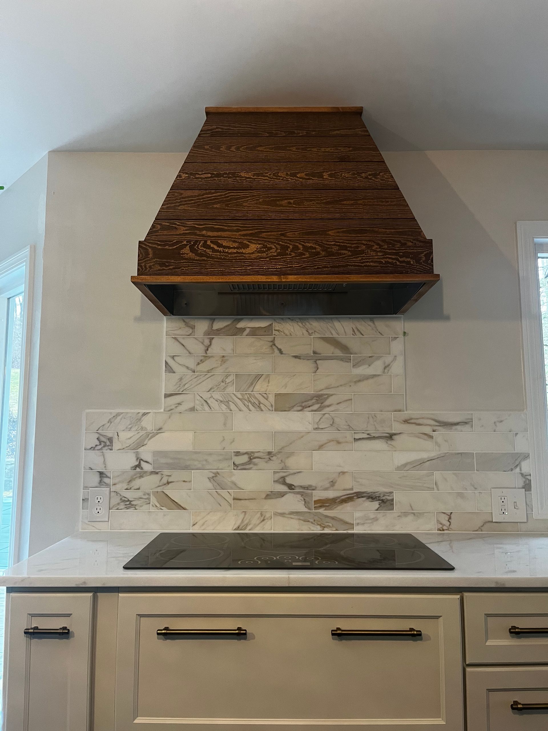 A kitchen stove area featuring a textured dark wood range hood above a marble-tiled backsplash and light-colored cabinetry.
