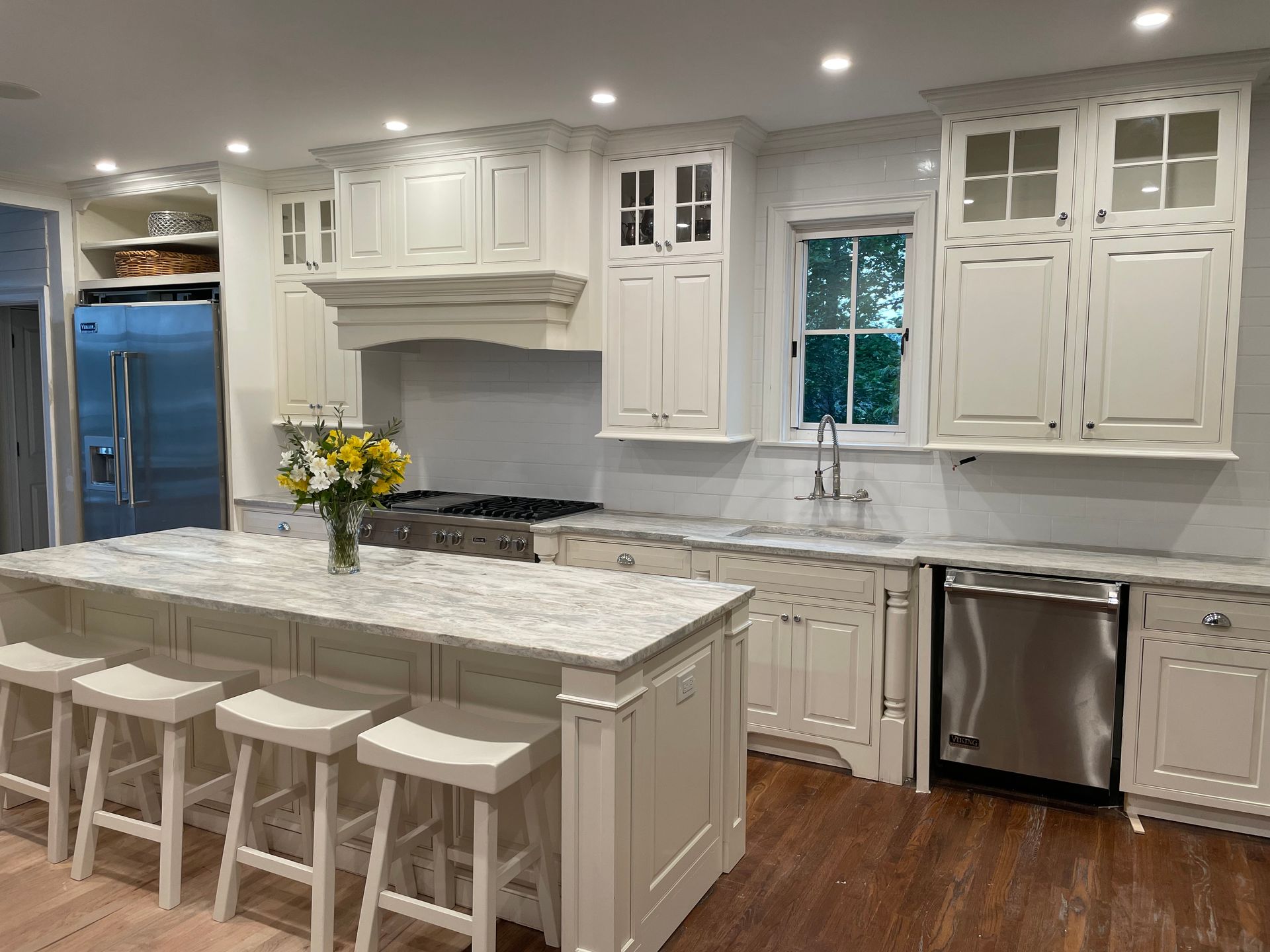 A kitchen featuring white cabinets, a marble-topped island with four stools, stainless steel appliances, and wood floors.