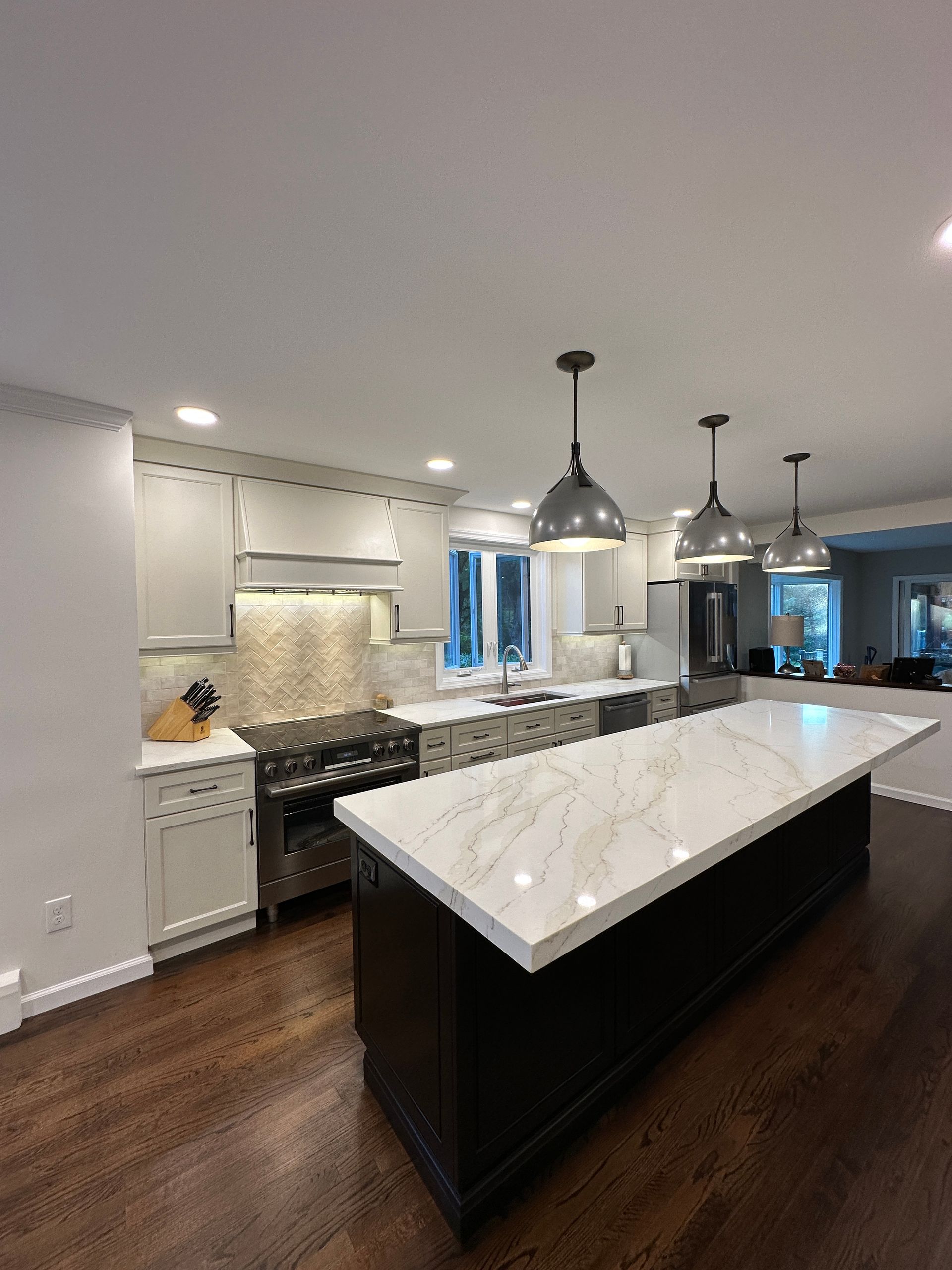 A modern kitchen featuring a dark island with white marble countertops, light-colored cabinets, and three hanging lights.