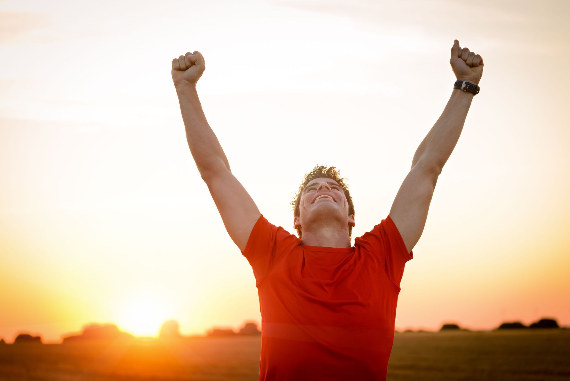 Man raises arms in celebration during a sunset in a field, wearing red shirt.