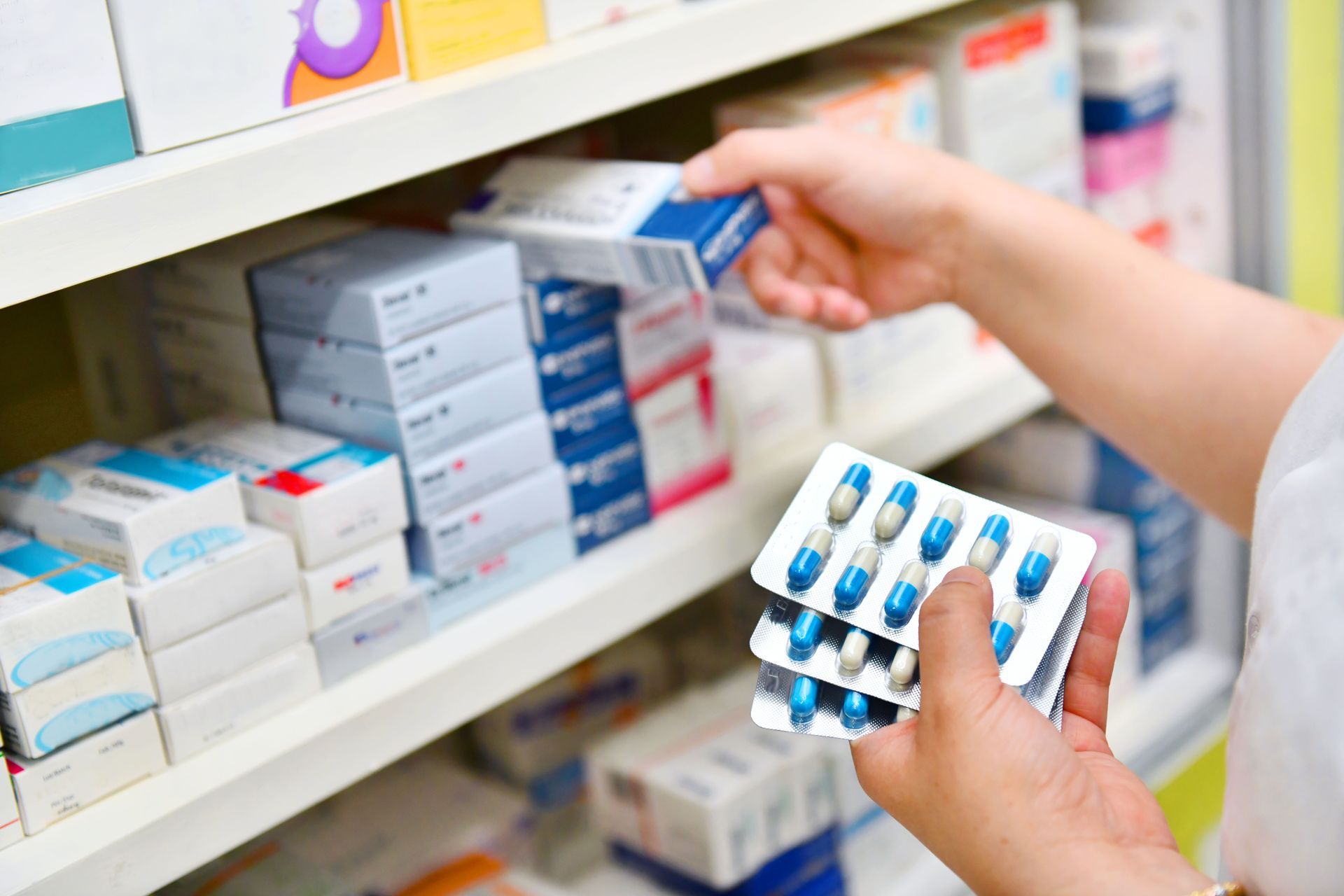 Person's hands holding medicine blister packs in a pharmacy, reaching for boxes on shelves.