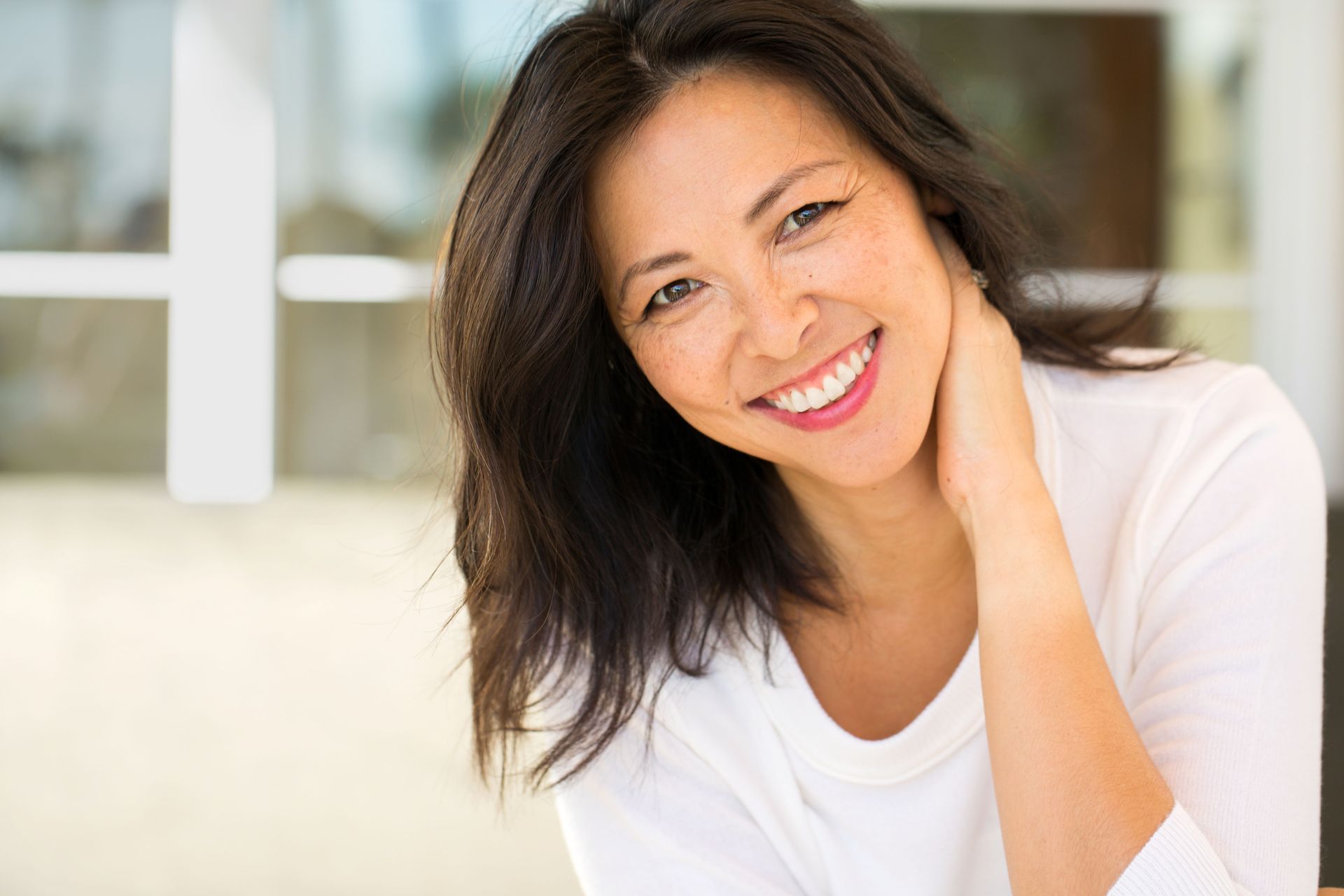 Woman with dark hair smiles, touches her neck, and looks at the camera. She's wearing a white shirt in a bright room.