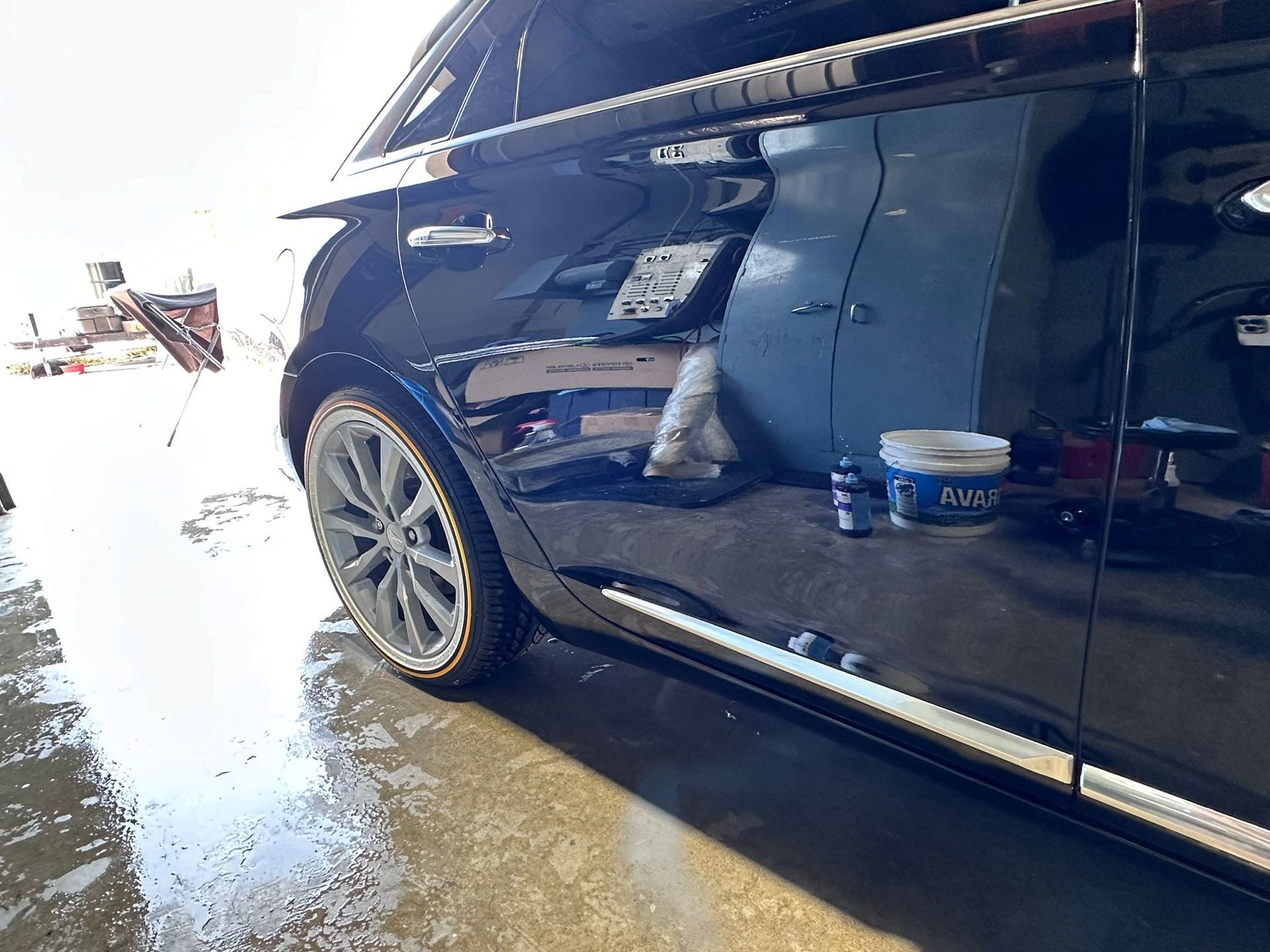 A black car is parked in a garage next to a bucket of wax.