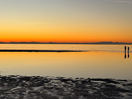 Sunset over calm water, two silhouetted figures on the shore. Orange and yellow sky reflects on the water.