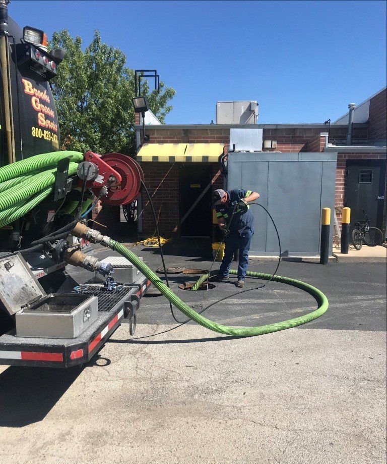A man operating a green hose attached to a black septic truck outside a building on a sunny day.