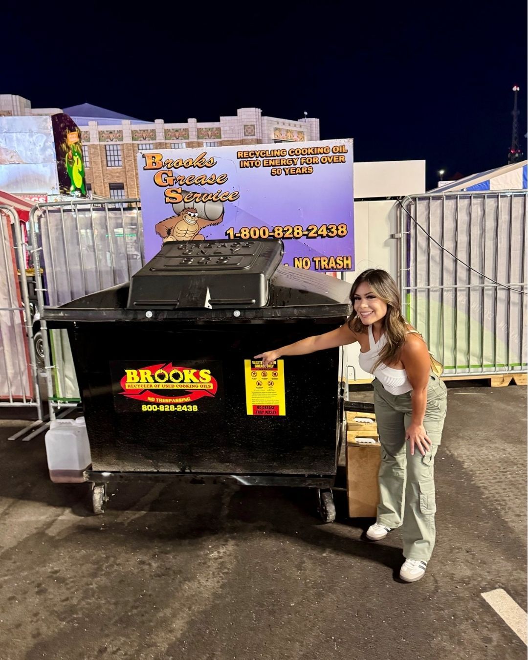 Woman points at a dumpster at night. Brooks Disposal logo, fair setting.