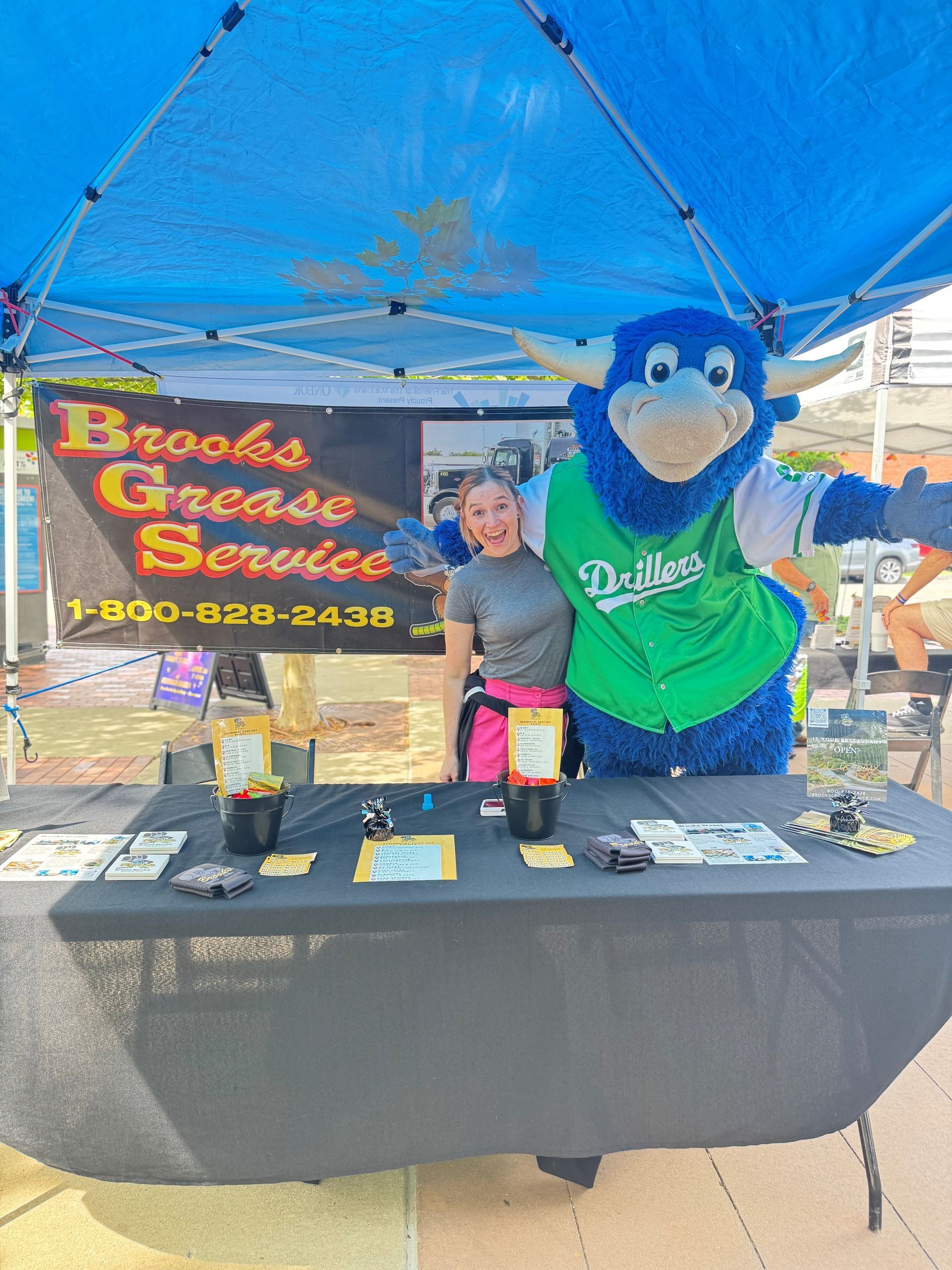A woman and a blue bull mascot at a Brooks Grease Service booth under a blue tent.