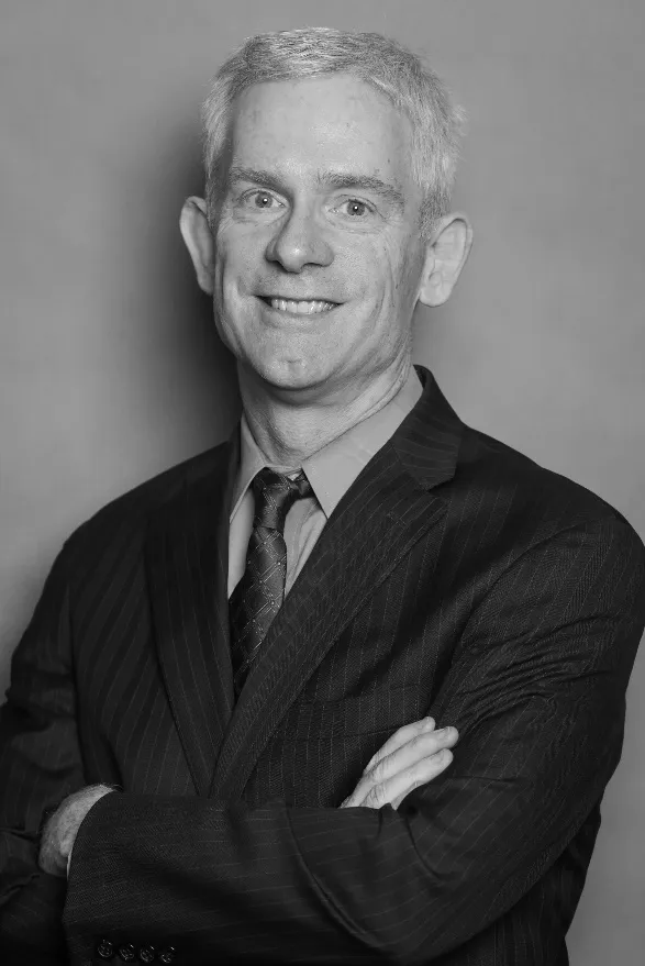 Portrait of a man in a dark suit with arms crossed, smiling against a plain gray background