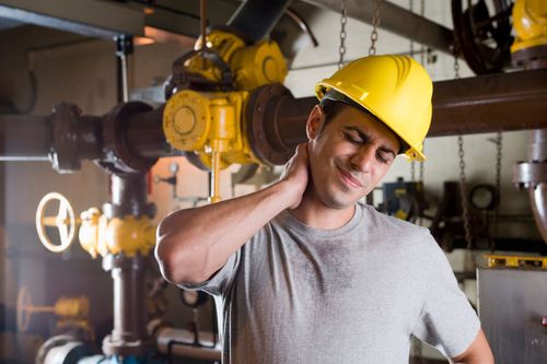 Worker in a yellow hard hat inspecting industrial pipes and valves in a factory setting.