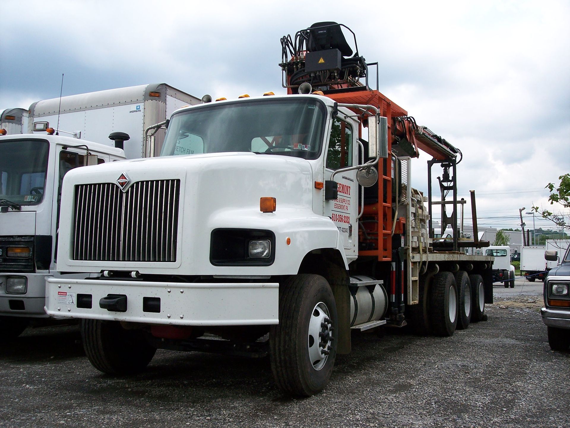 A white International truck equipped with a crane and flatbed parked on a gravel lot under an overcast sky.