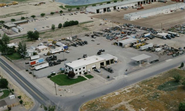 An aerial view of a industrial facility with a central white building, outdoor storage, a parking lot, and access roads.