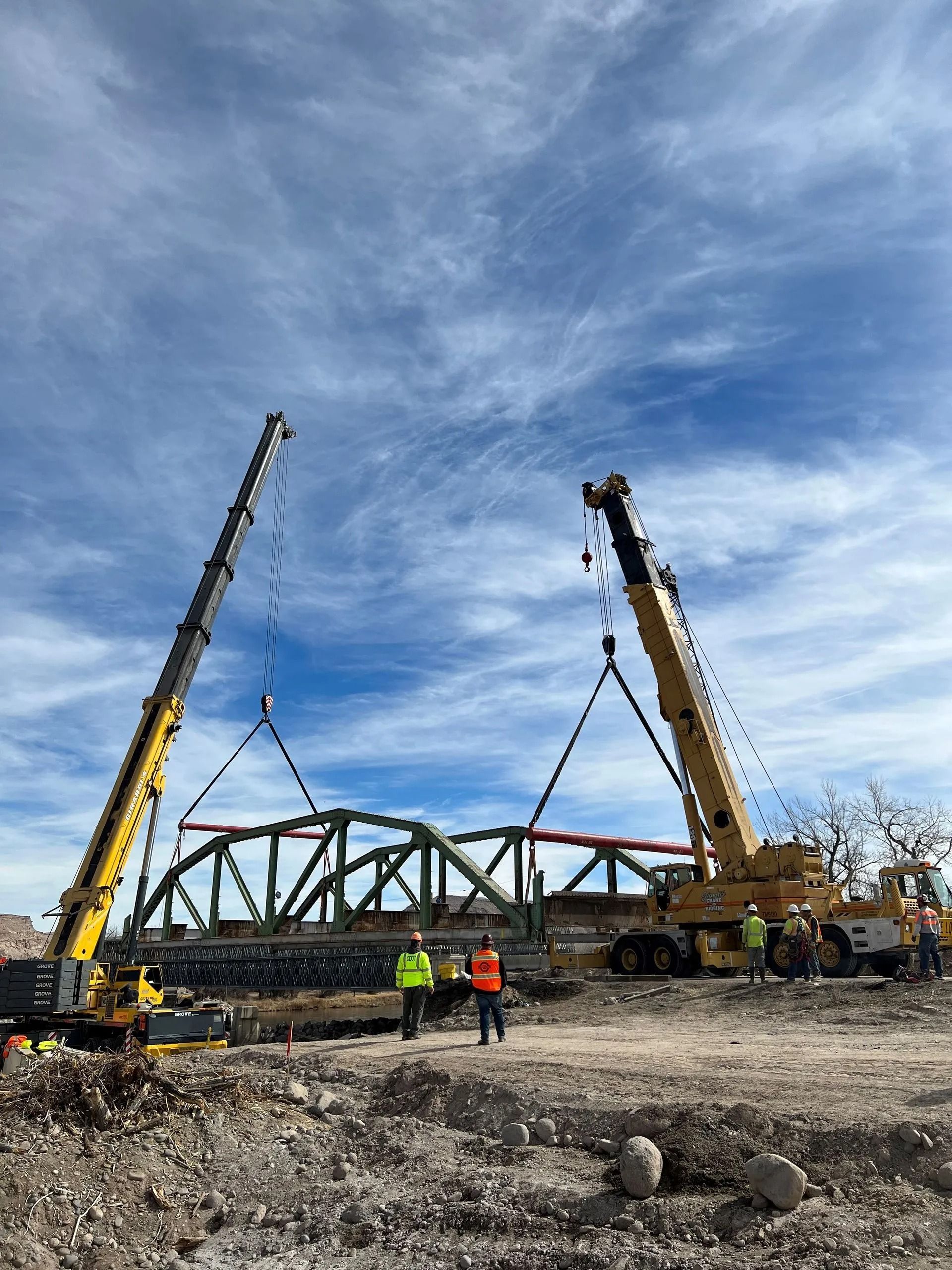 Two cranes lift a green steel bridge truss under a blue sky, while workers in safety vests stand on the ground nearby.