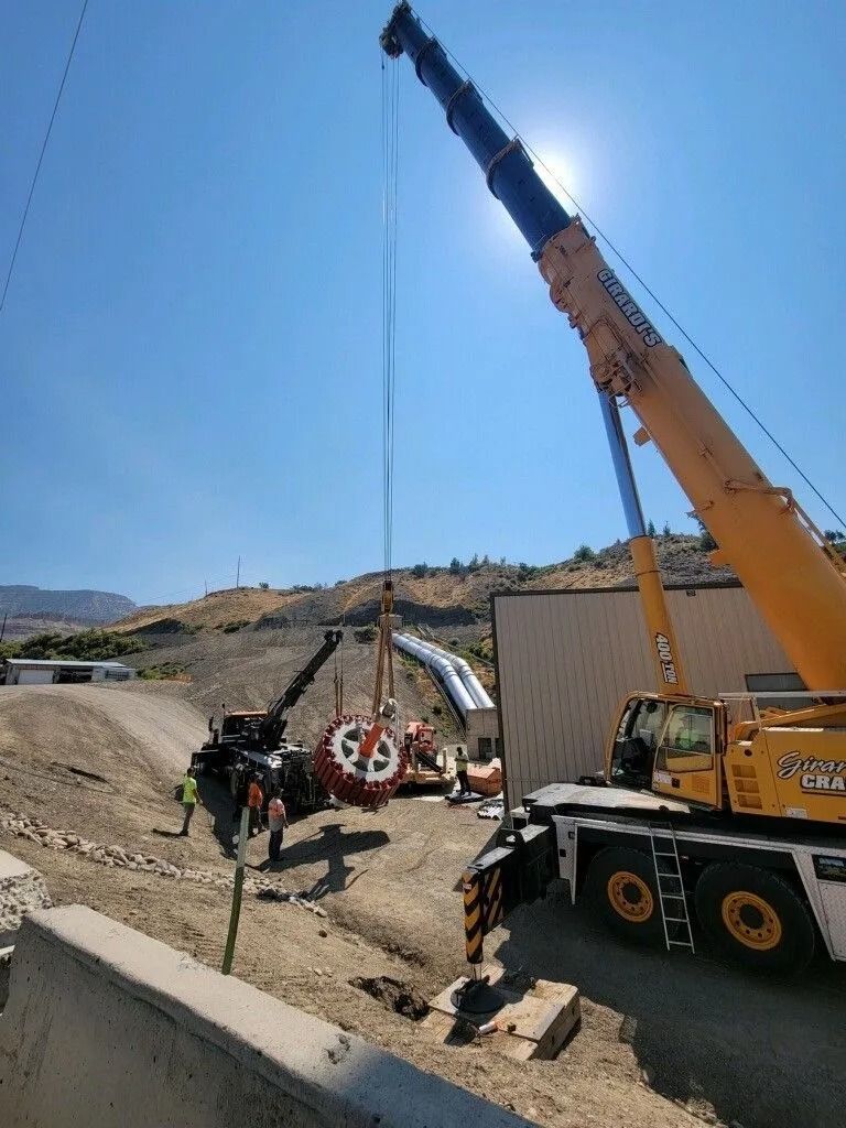 Crane lifting heavy equipment at a construction site with workers nearby under a clear sky