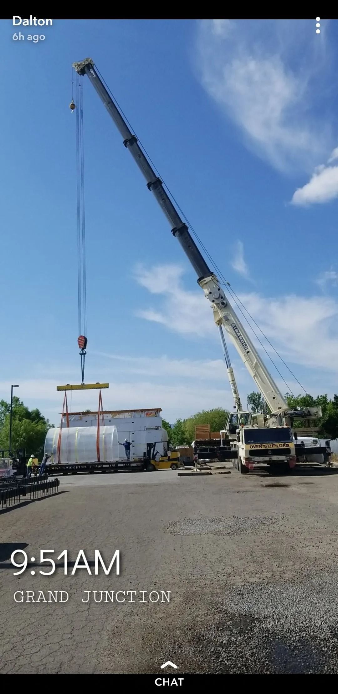 A large mobile crane lifts a white, cylindrical industrial tank outdoors under a clear blue sky.