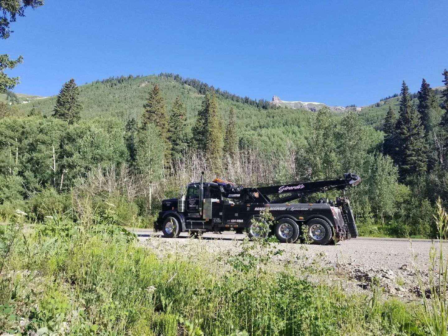 A black heavy-duty tow truck parked on a gravel roadside in front of a mountain covered with trees.