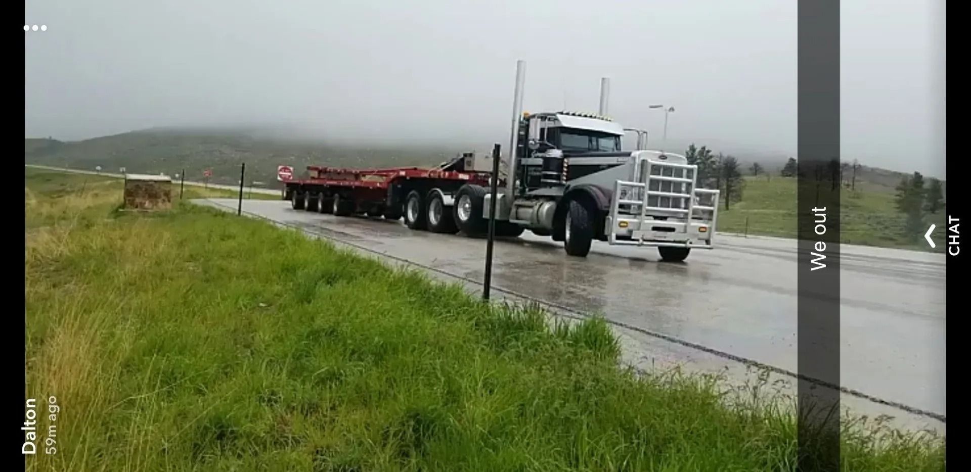 A semi-truck with a large metal grille guard pulls an empty flatbed trailer down a wet road near a grassy hillside.