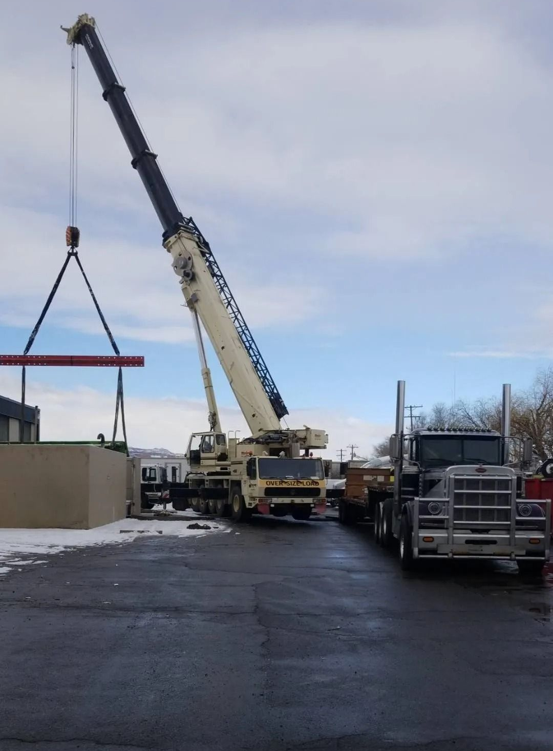 A large crane lifting a long red pipe at a snowy outdoor site near a parked semi-truck.