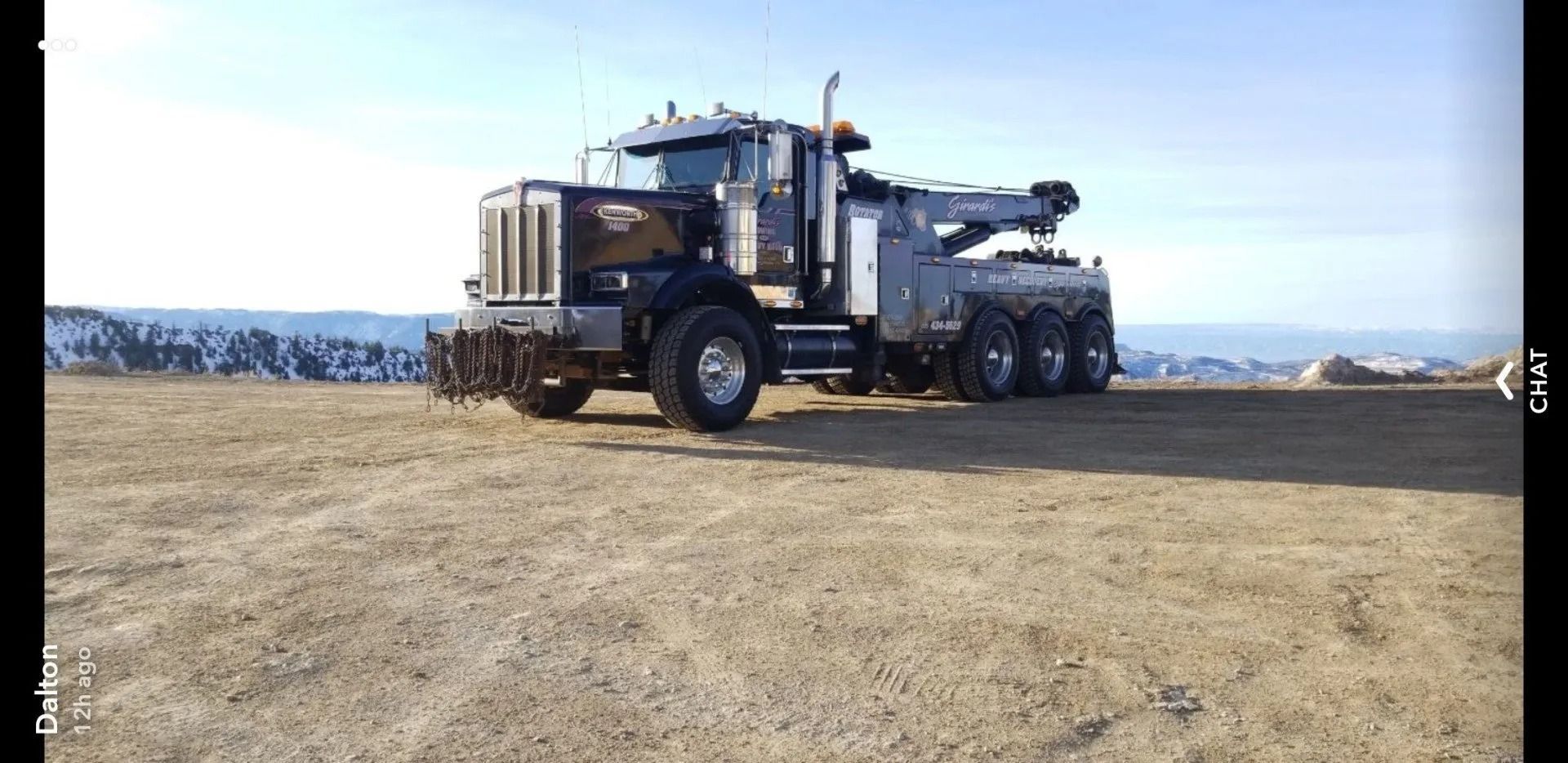 A large, heavy-duty black and silver tow truck sits parked in an open, dry, grassy field under a bright sky.