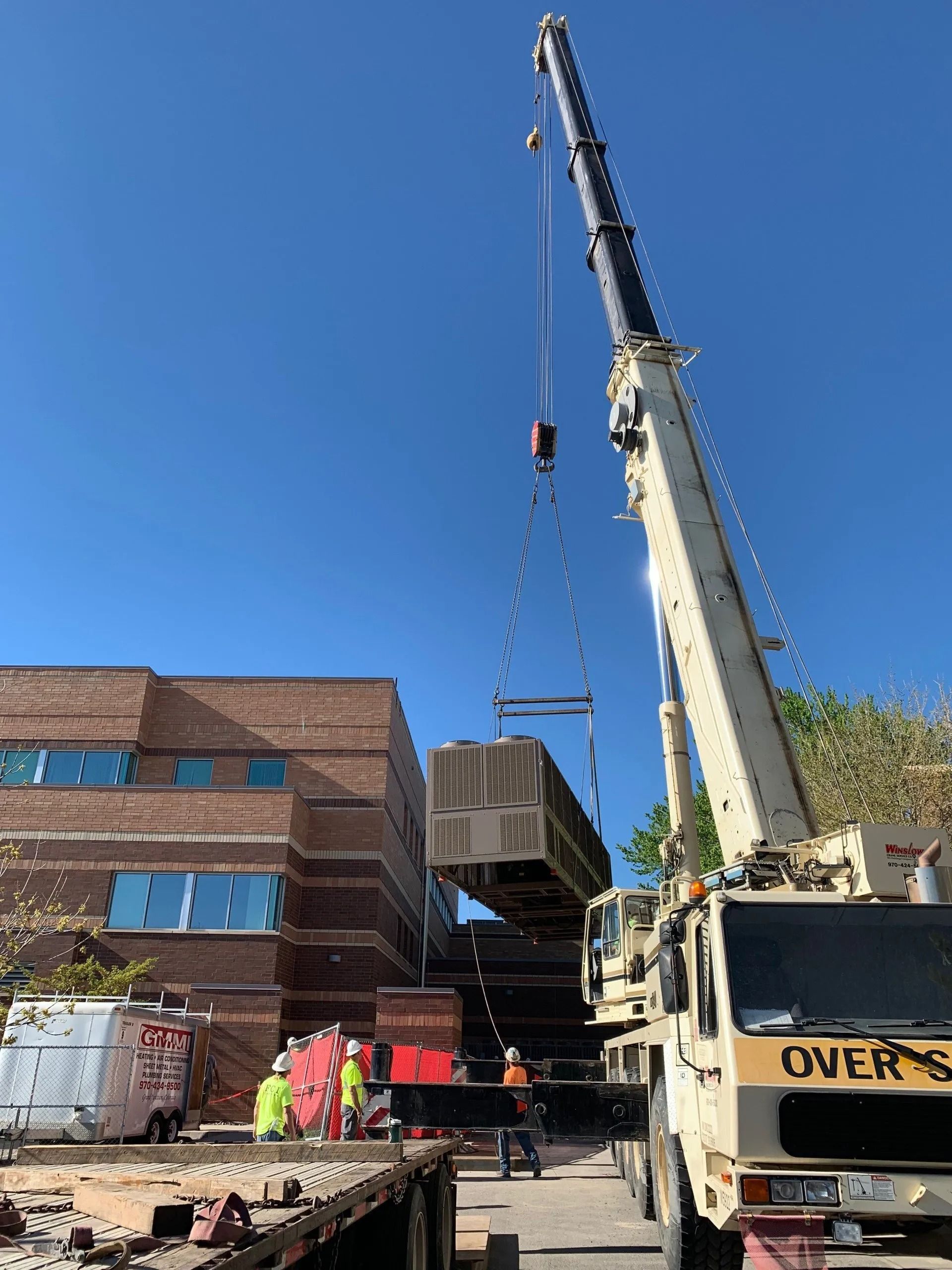 A crane hoists a large HVAC unit near a brick building while construction workers stand nearby under a clear blue sky.