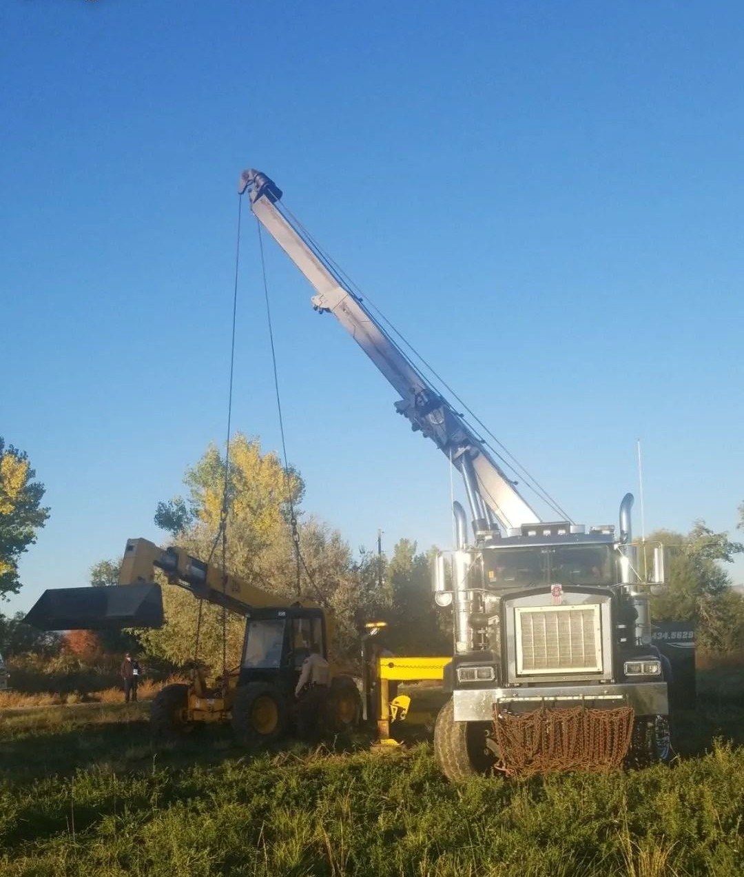 A large mobile crane lifts a yellow agricultural loader in a field under a clear blue sky.