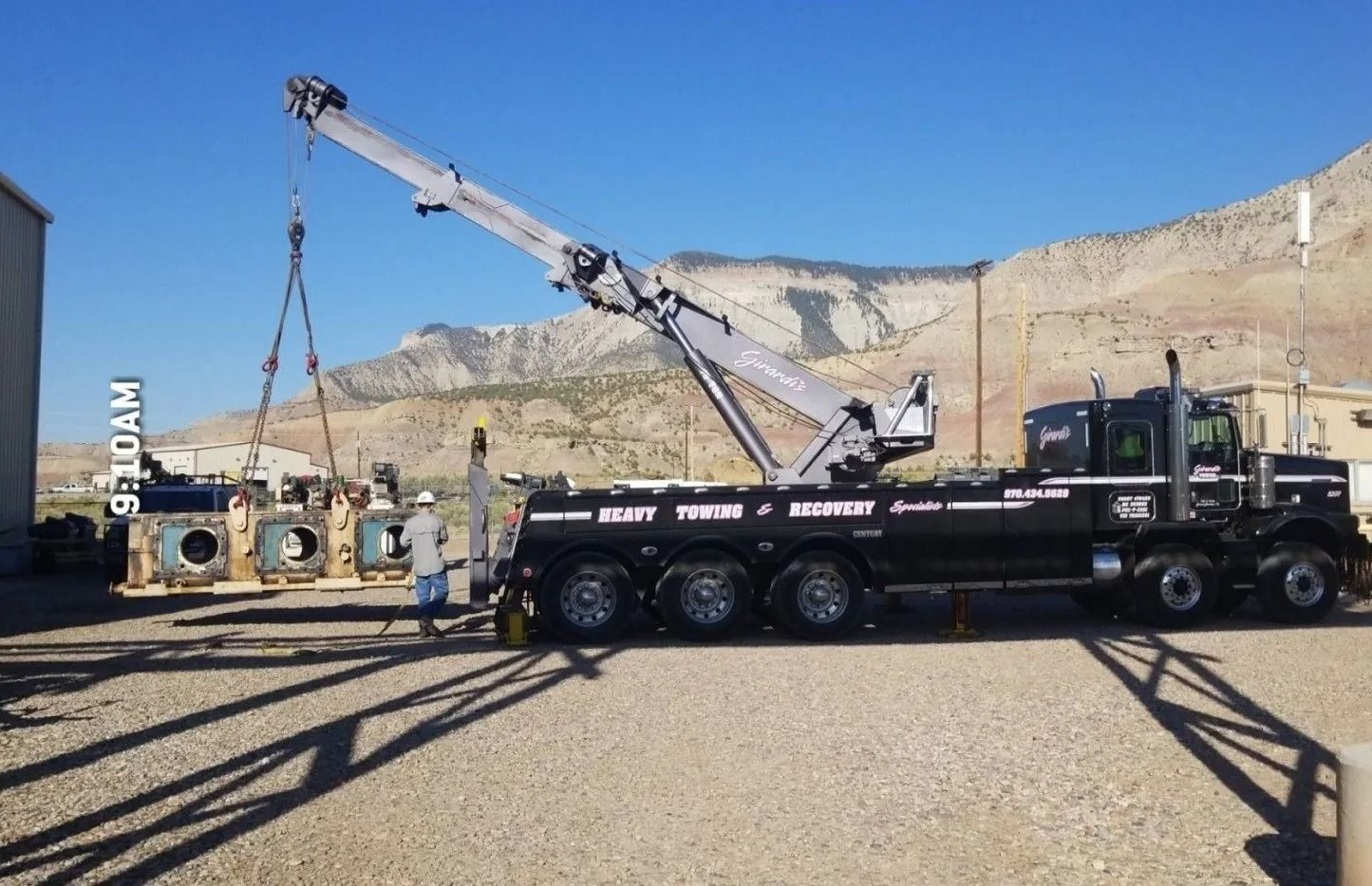 A large black heavy-duty crane truck lifts a mechanical engine part outdoors against a desert landscape and mountains.