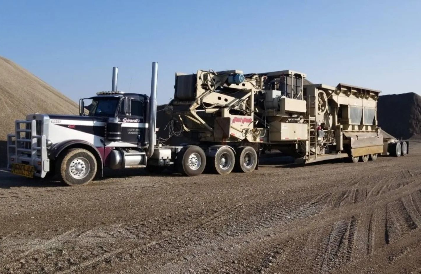 A semi-truck towing a large, industrial rock-crushing machine across a gravel lot on a clear day.