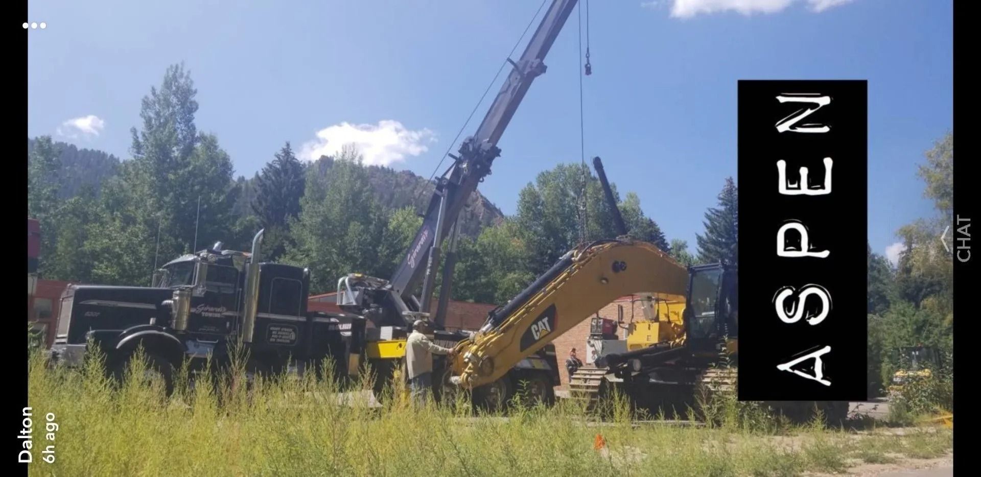 A crane lifts a yellow Caterpillar excavator in a sunny, grassy area with mountains in the background, labeled 
